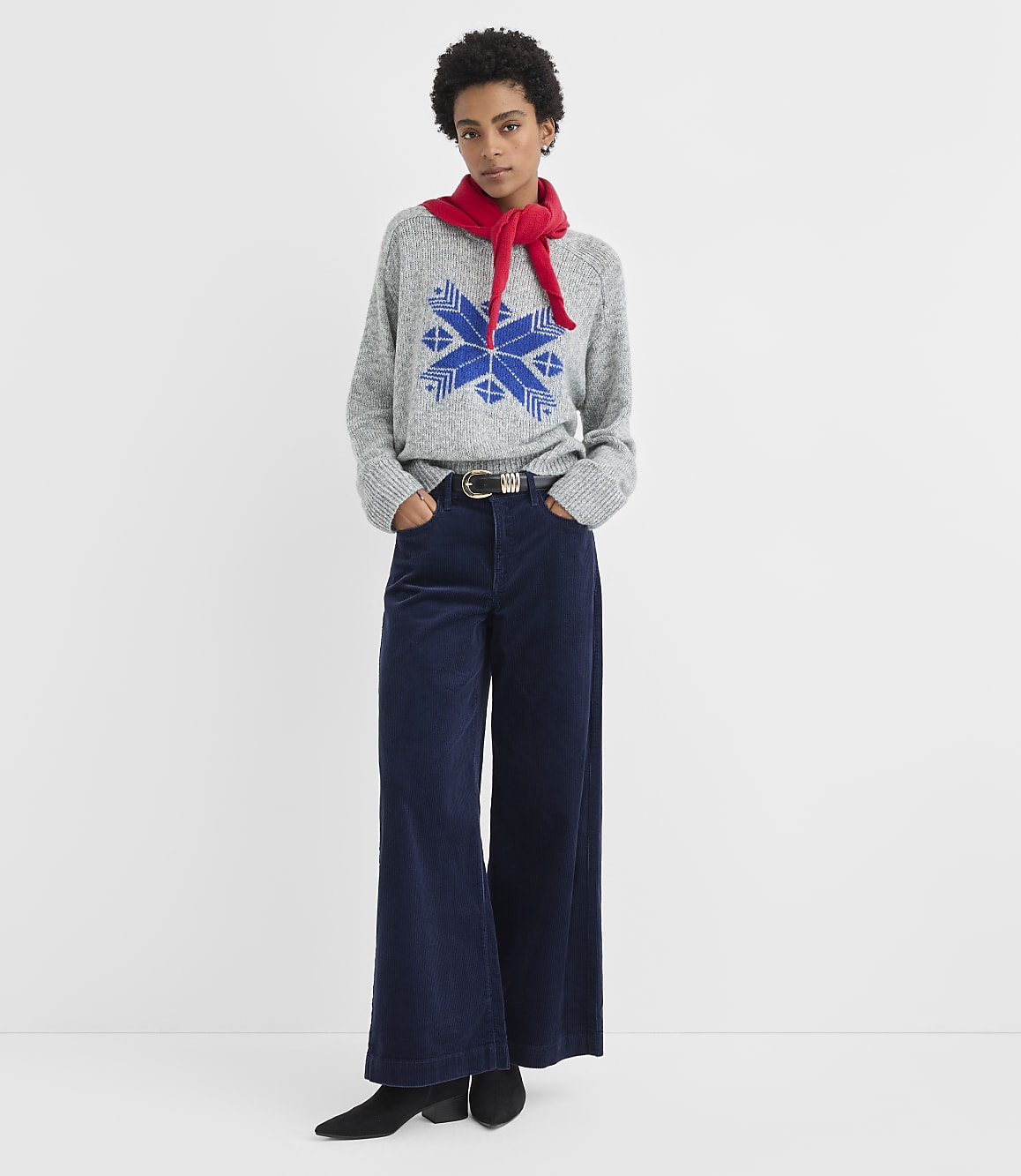 A woman wearing a grey patterned sweater, red scarf, and wide-leg navy trousers stands against a plain white background.