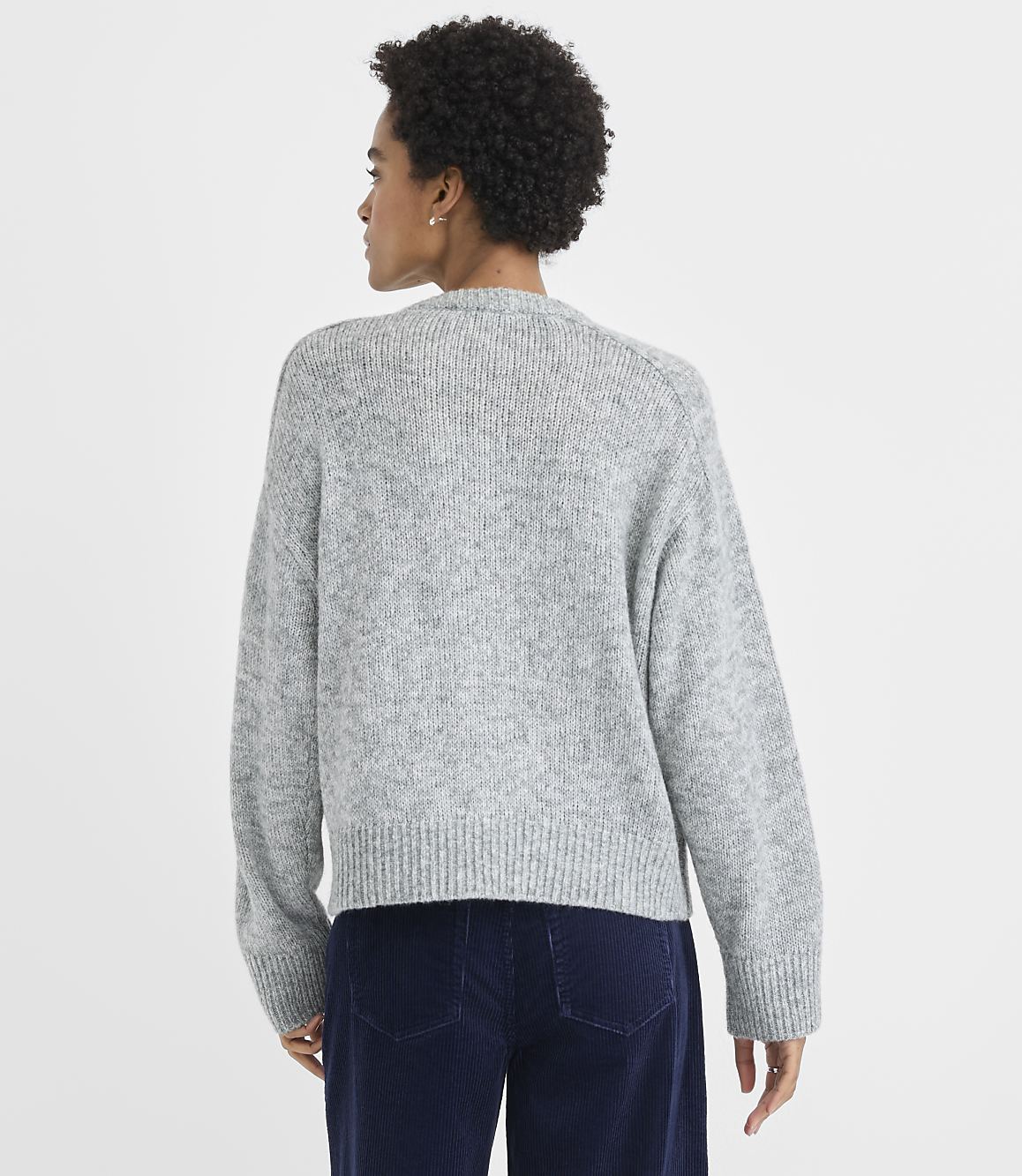 A woman wearing a grey knit sweater and navy blue corduroy pants stands against a plain white background.