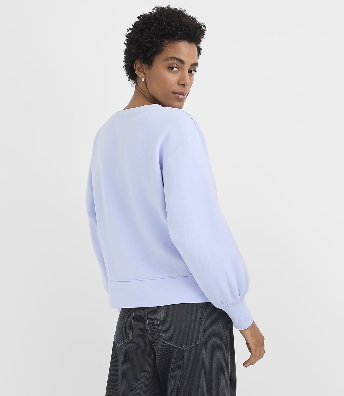 A woman wearing a light blue sweatshirt and dark grey trousers stands against a plain white background.
