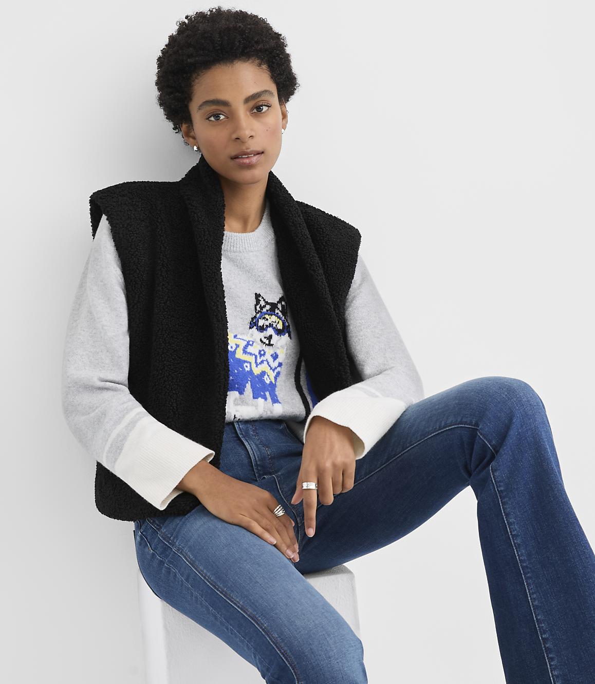 A woman wearing a grey patterned sweater, black vest, and blue jeans sits on a white stool against a plain white background.