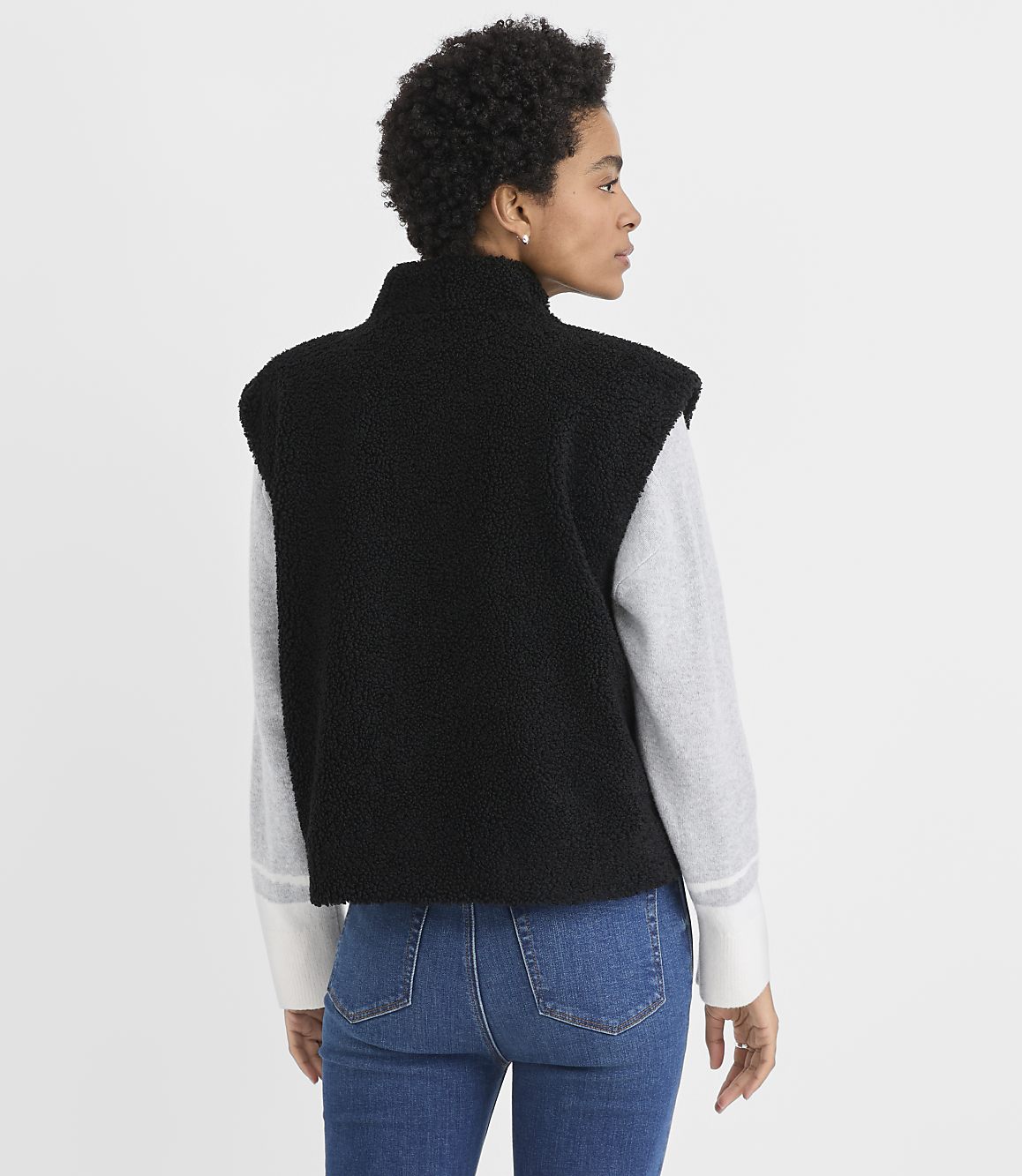 A woman wearing a black fleece vest over a grey long-sleeved shirt and blue jeans stands against a plain white background.