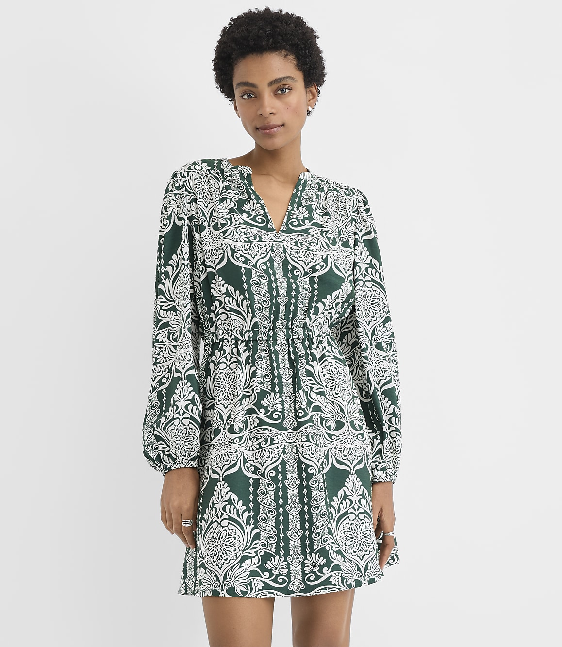 A woman wearing a green and white patterned mini dress stands against a plain white background.