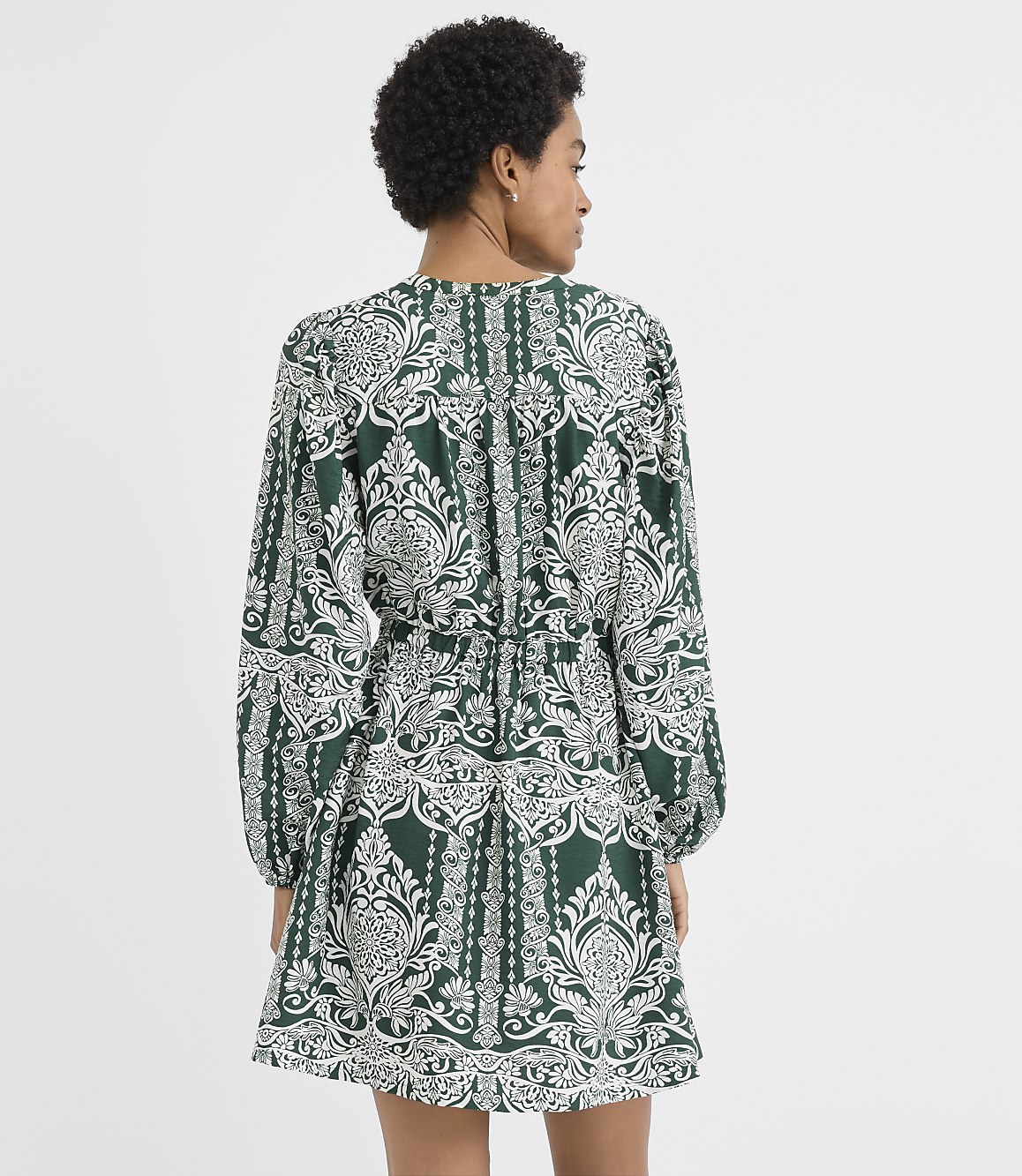 A woman wearing a green and white patterned dress stands with her back to the camera against a plain white background.