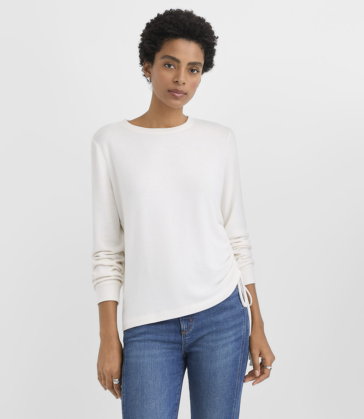 A woman wearing a white long-sleeved shirt and blue jeans stands against a plain white background.