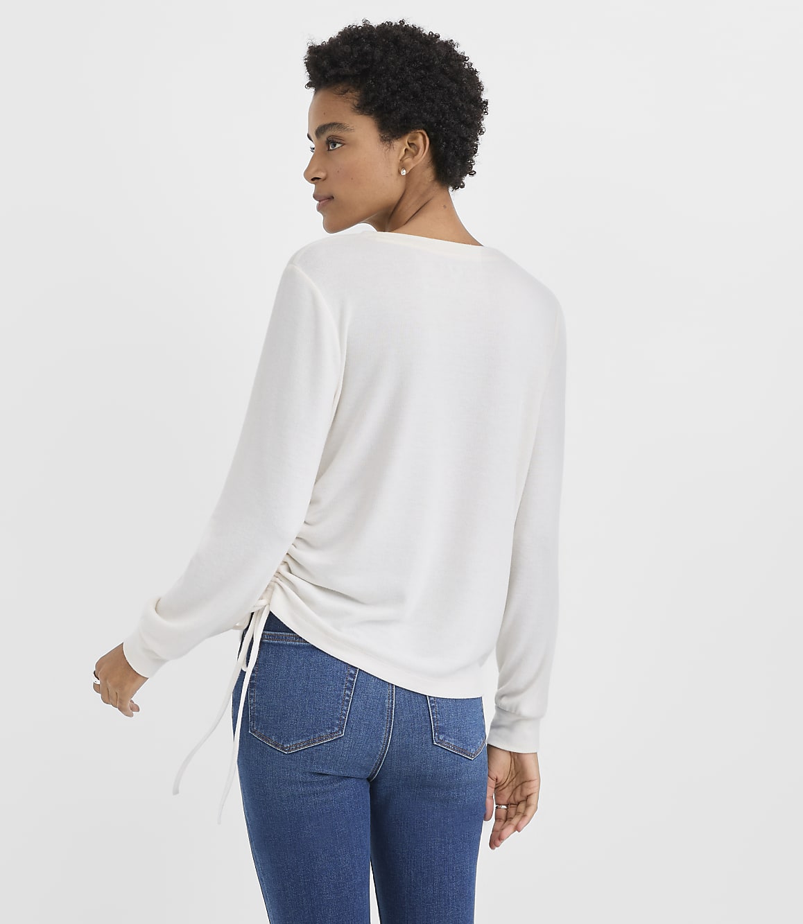 A woman wearing a white long-sleeved top and blue jeans stands against a plain white background.