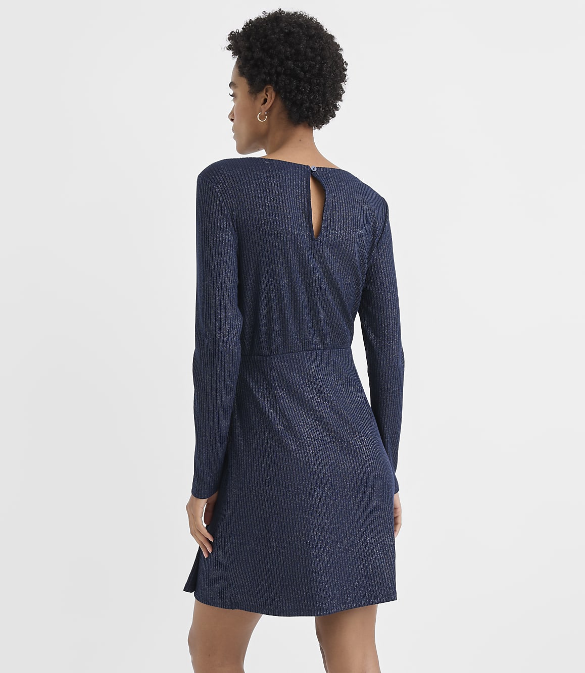 A woman with curly hair wearing a long-sleeved navy blue ribbed dress stands against a plain white background.