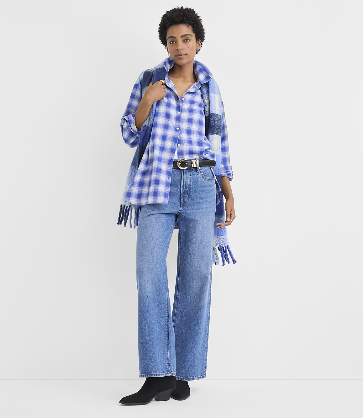 A woman wearing a blue plaid shirt, wide-leg jeans, and black boots stands against a plain white background.