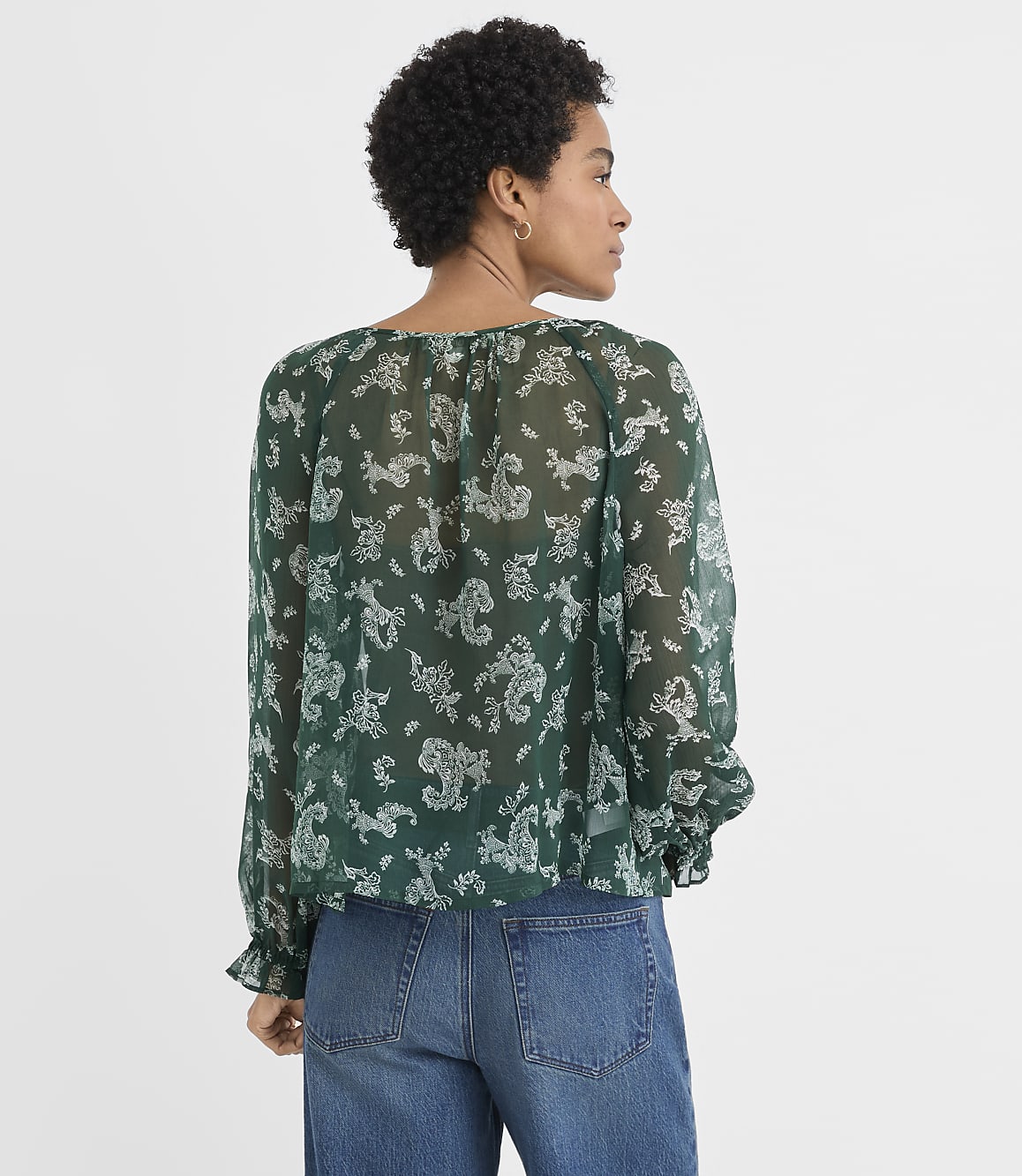 A woman wearing a green floral print blouse and blue jeans stands against a plain white background.