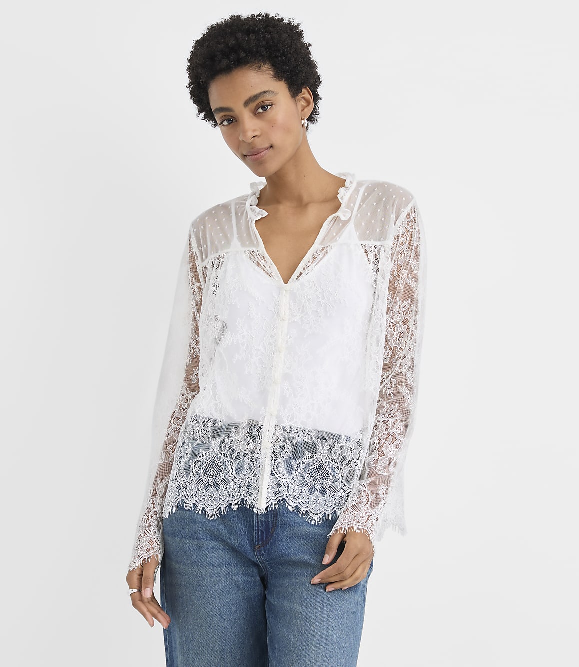 A woman wearing a white lace blouse and blue jeans stands against a plain white background.