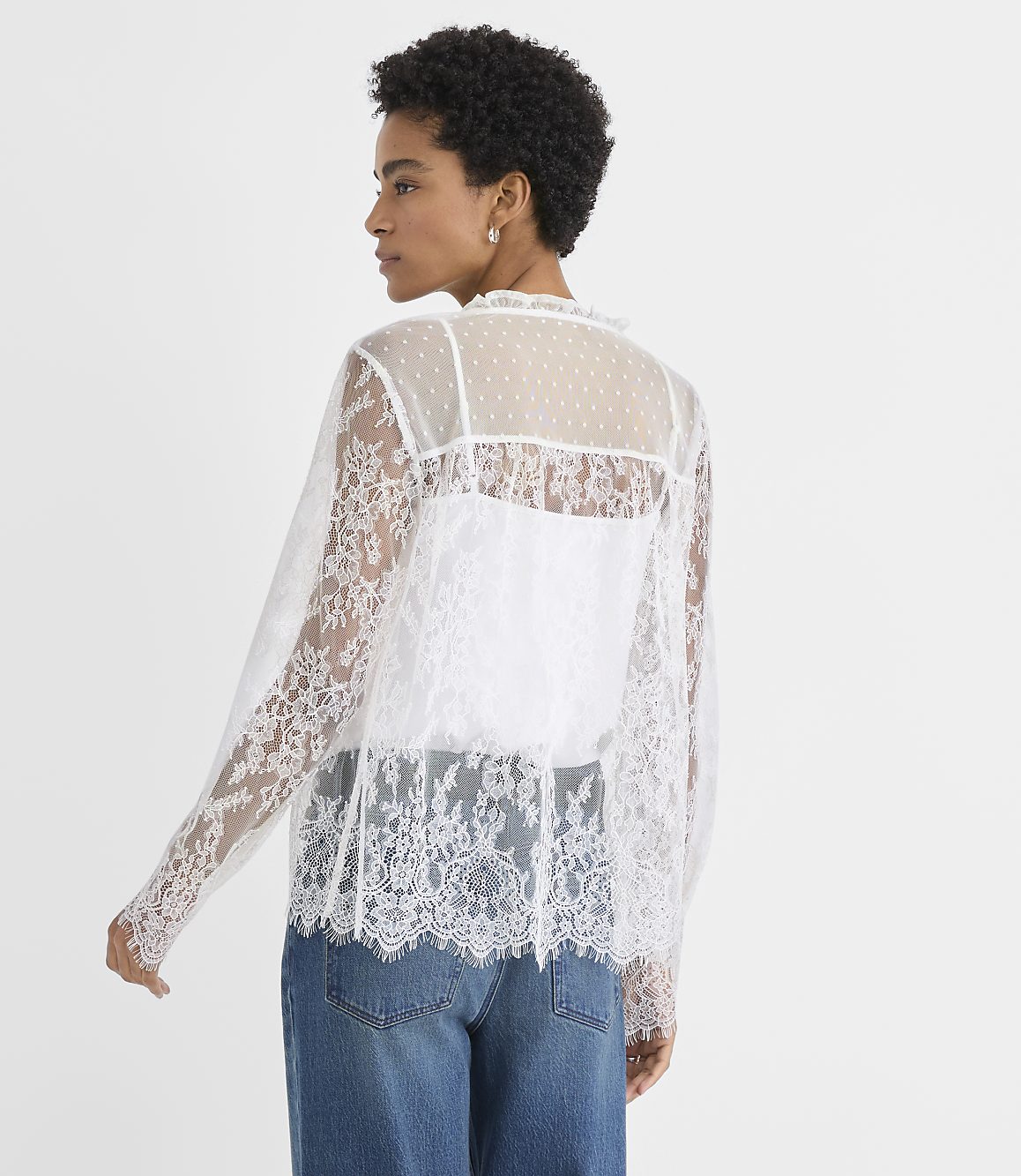 A woman wearing a white lace top and blue jeans stands against a plain white background.