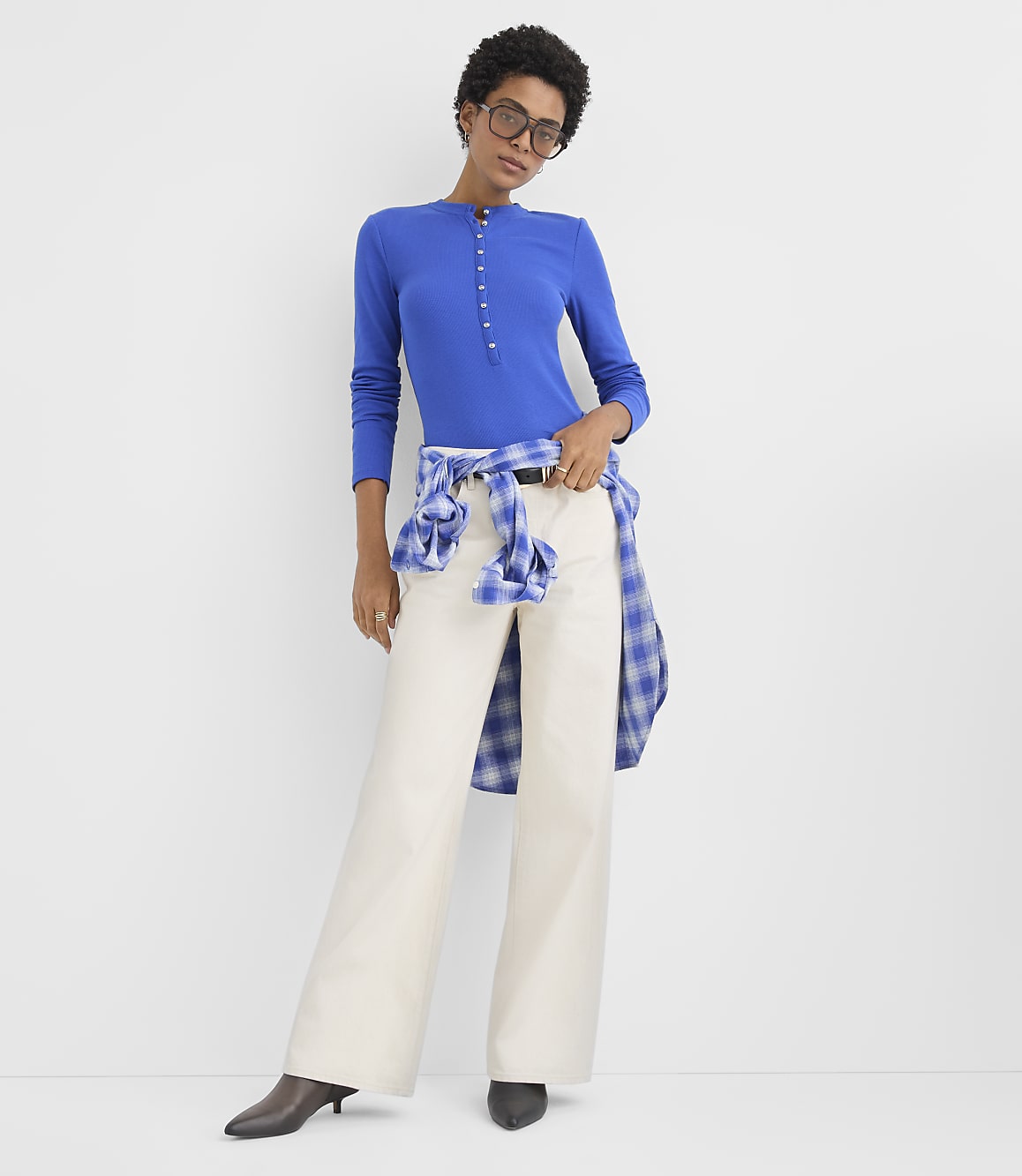 A woman wearing a blue long-sleeved top and wide-leg white pants stands against a plain white background.