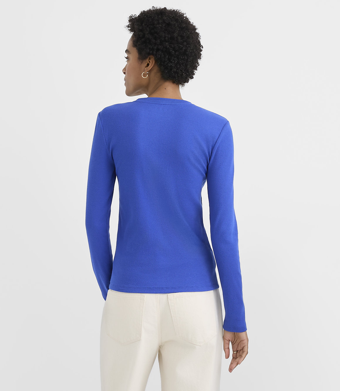 A woman wearing a bright blue long-sleeved shirt and white pants stands with her back to the camera against a plain white background.