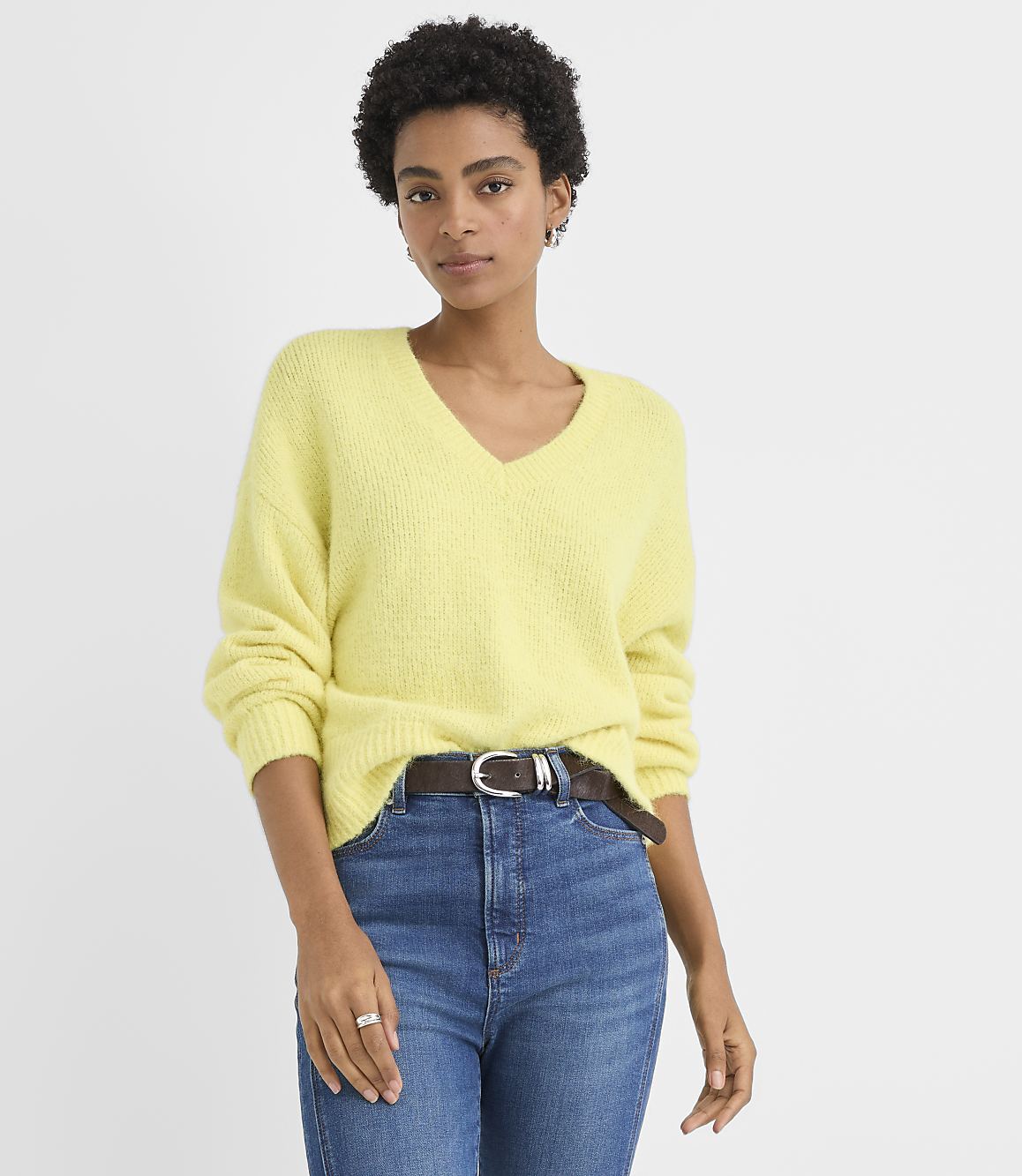 A woman wearing a yellow V-neck sweater and blue jeans stands against a plain white background.