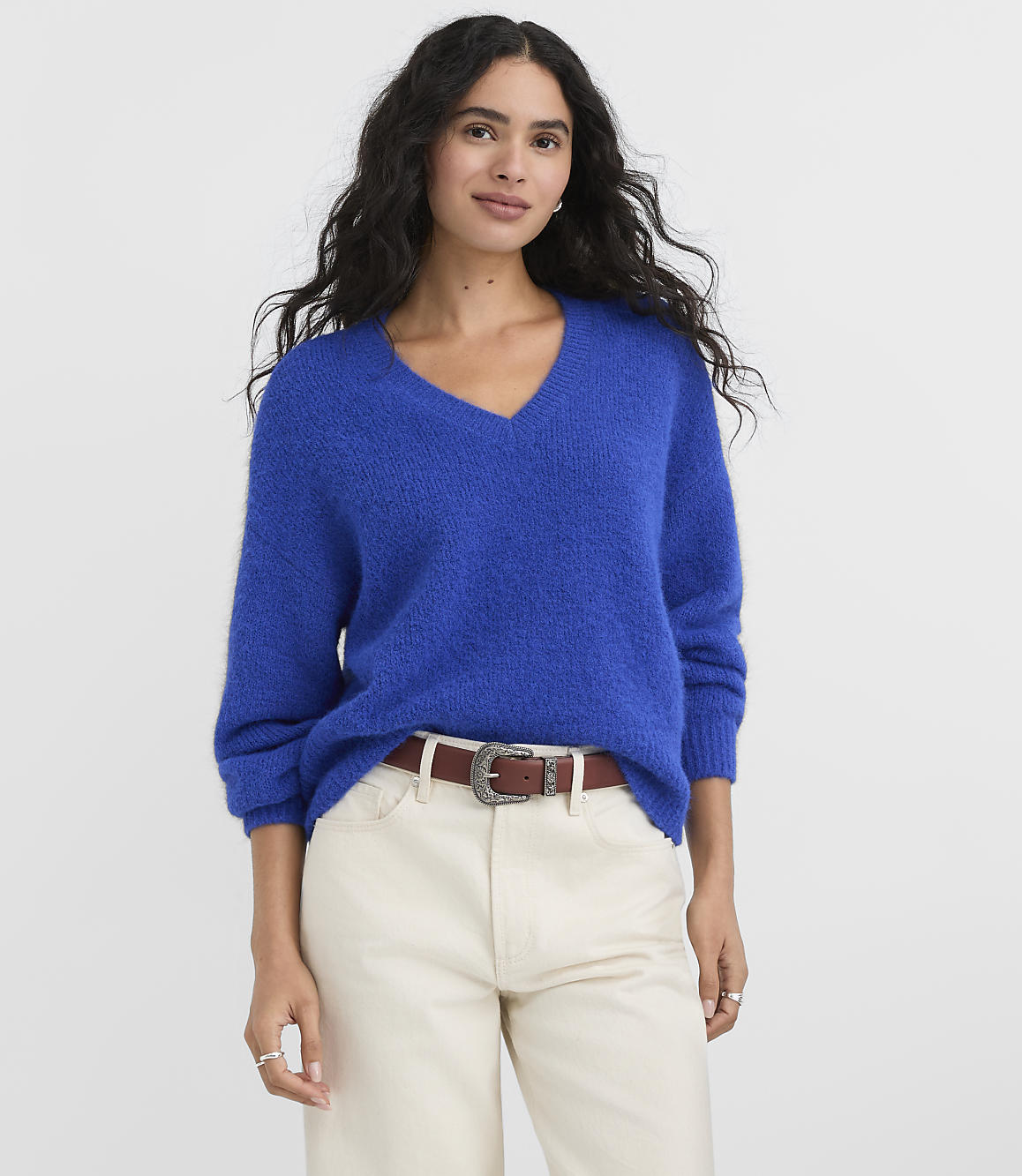 A woman wearing a bright blue v-neck sweater and cream-colored pants stands against a plain white background.