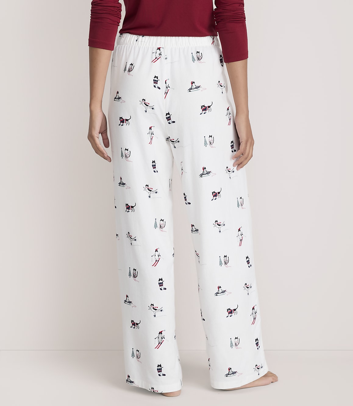 A person wears a red long-sleeved shirt and white pajama pants featuring a festive dog print, standing against a plain, light-colored background.