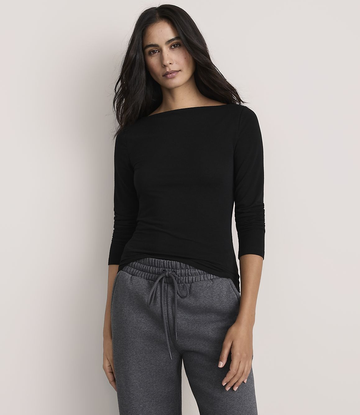 A woman wearing a black long-sleeved shirt and grey sweatpants stands against a plain, light-colored background.
