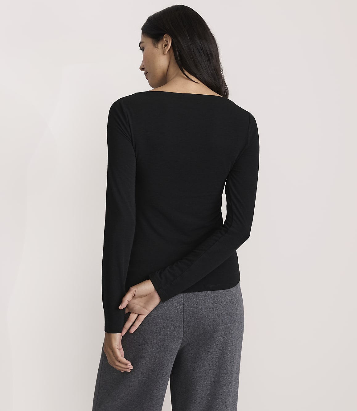 A woman wearing a black long-sleeved shirt and grey pants stands with her back to the camera against a plain white background.