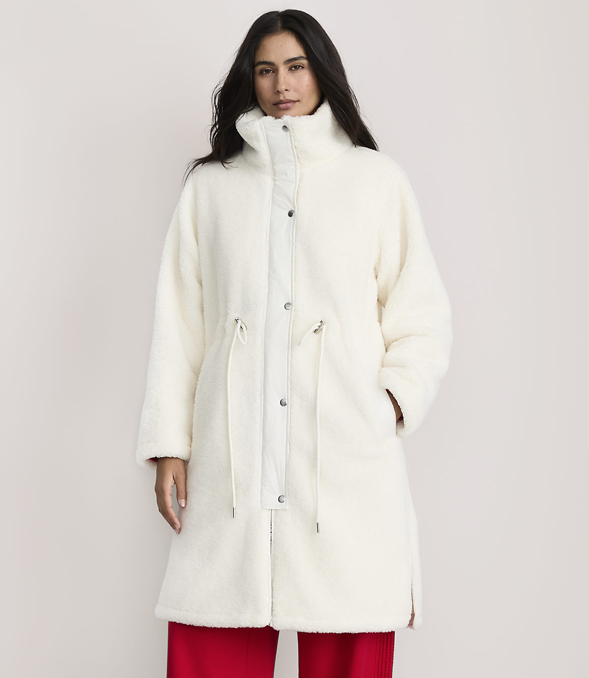 A woman wearing a long, white fuzzy coat and red pants stands against a plain, light-colored background.