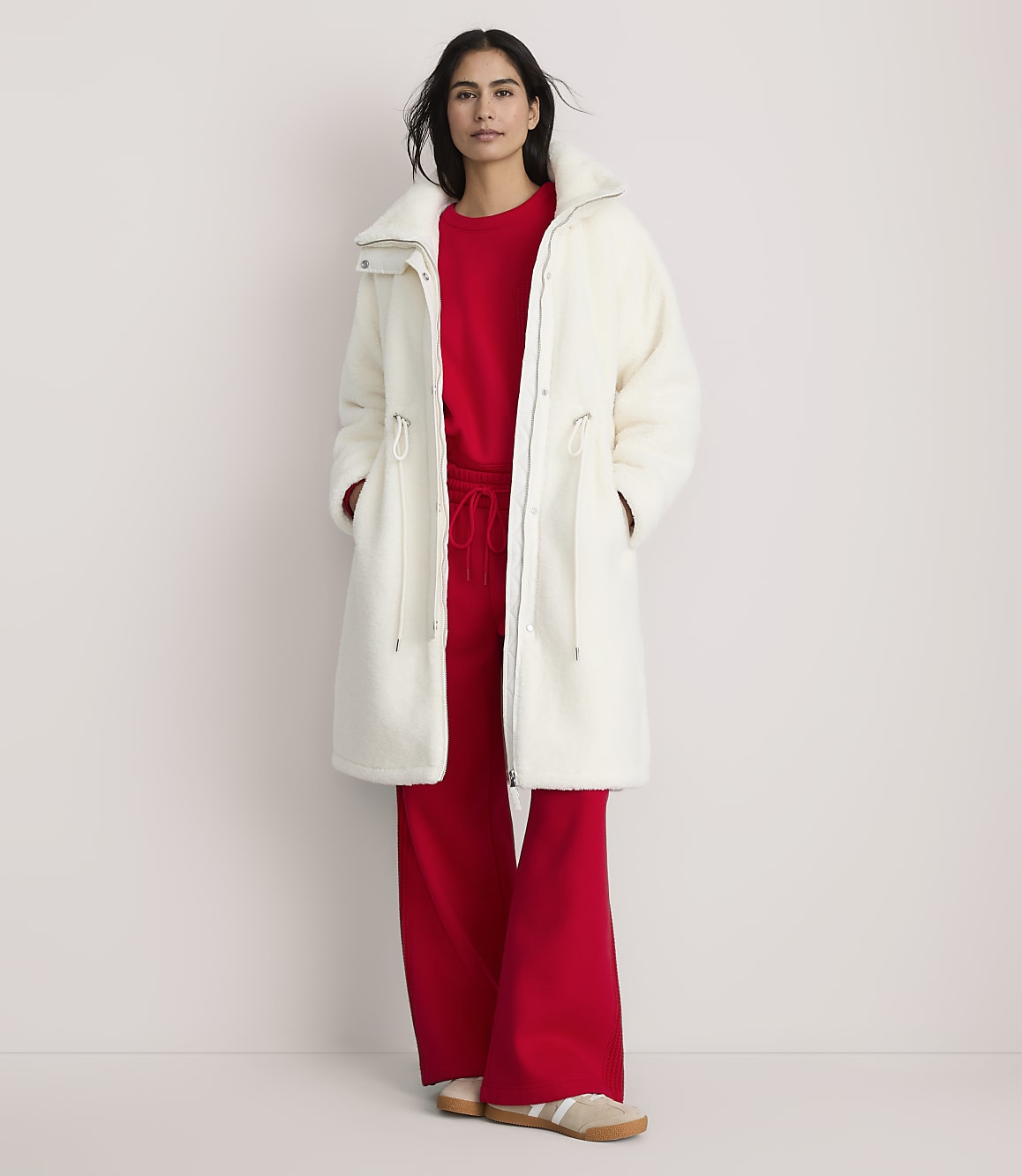 A woman wearing a long white fuzzy coat over a red outfit stands against a plain, light-colored background.