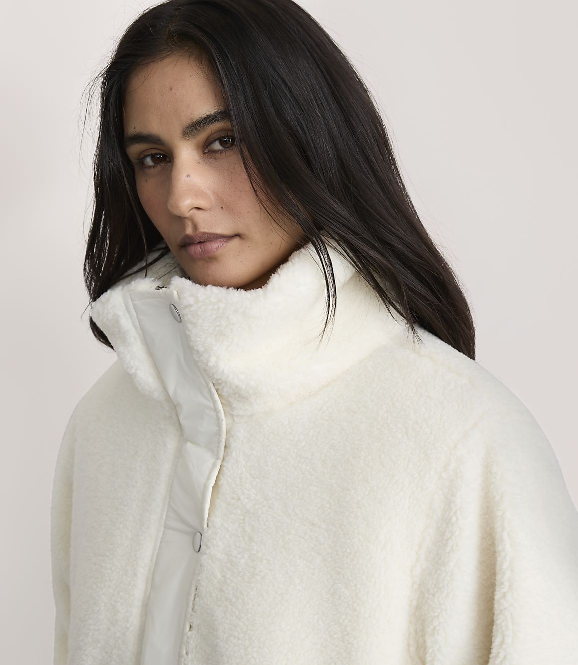 A woman with long dark hair wearing a white fuzzy jacket stands against a plain, light-colored background.