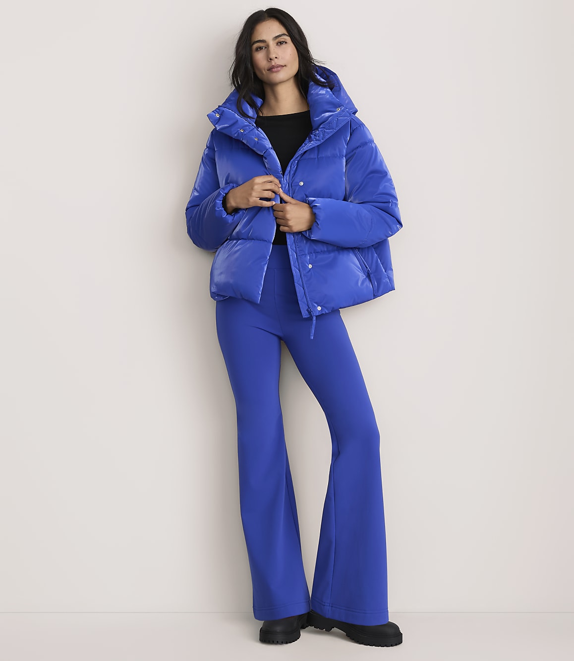 A woman wearing a bright blue puffer jacket and matching flared trousers stands against a plain, light-colored background.