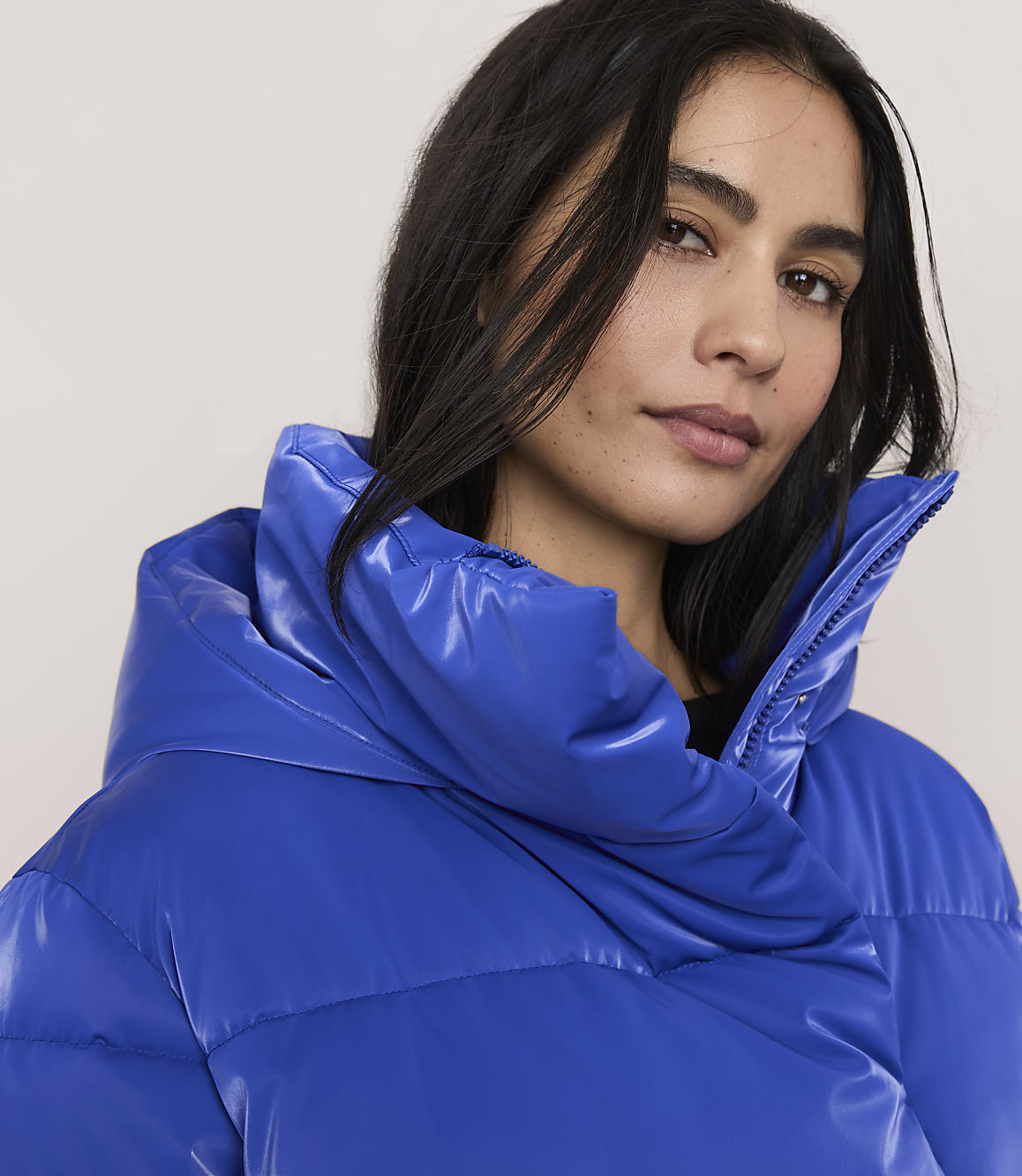 A woman with dark hair wearing a bright blue puffer jacket poses against a plain, light-colored background.
