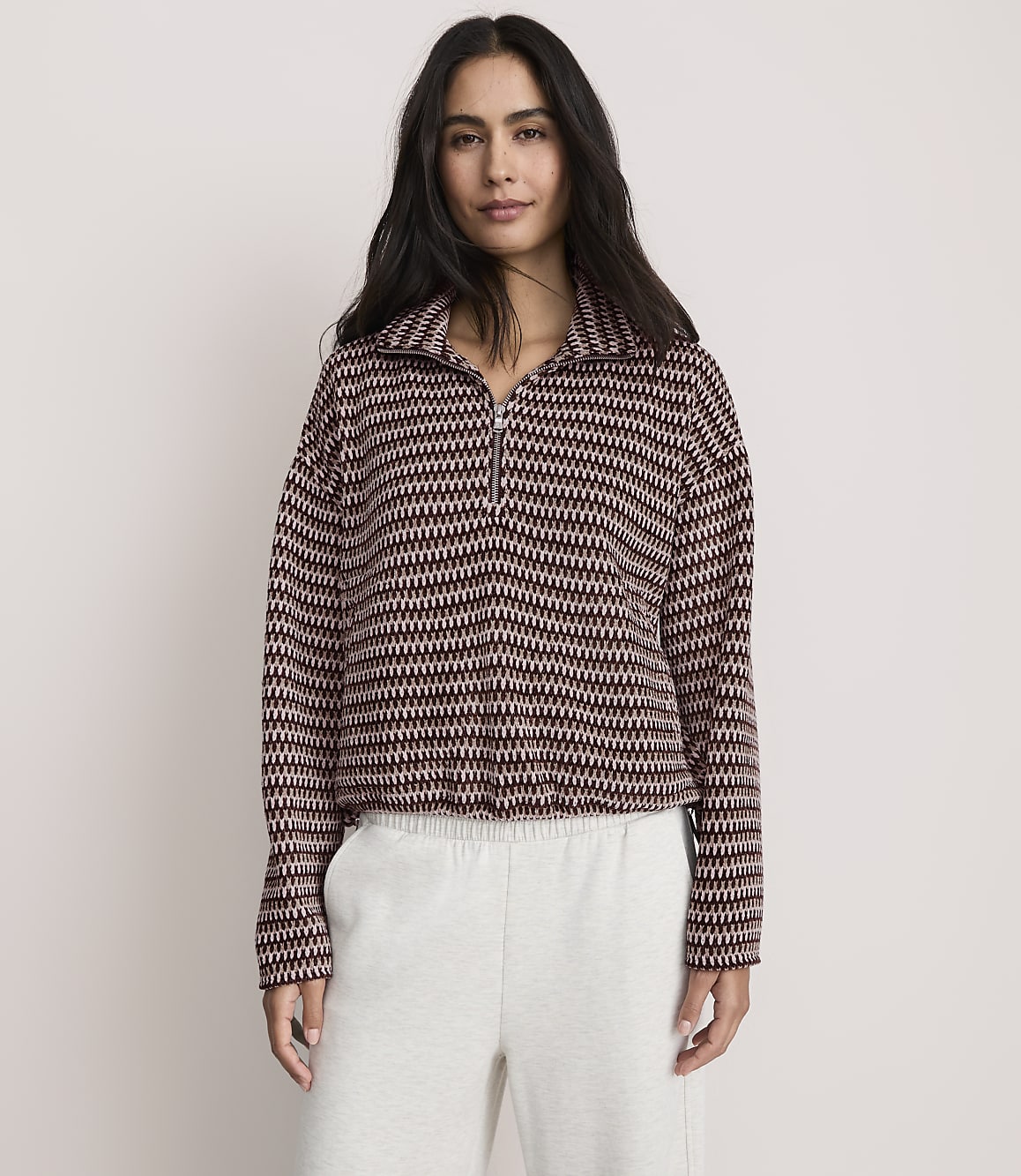 A woman wearing a patterned maroon and white quarter-zip pullover and light grey sweatpants stands against a plain, light-colored background.