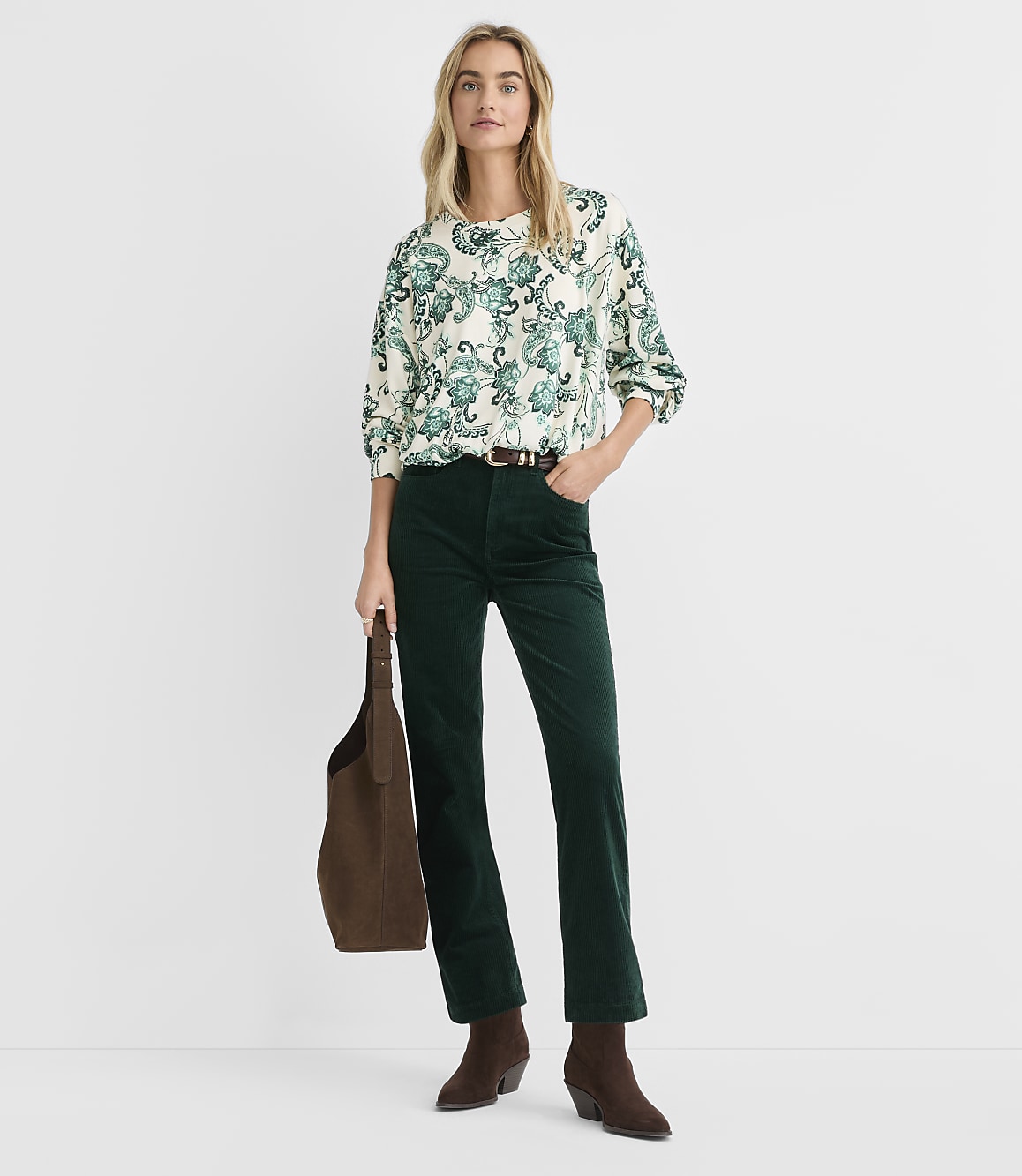 A woman wearing a patterned blouse and dark green corduroy pants stands against a plain white background.