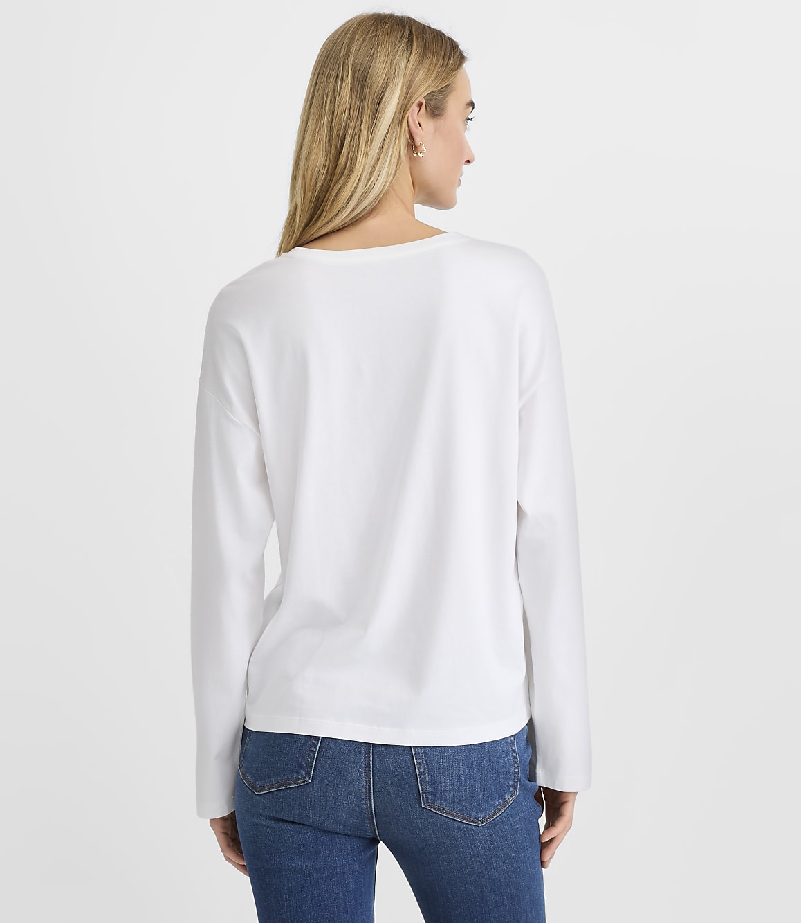 A woman wearing a white long-sleeved shirt and blue jeans stands with her back to the camera against a plain white background.