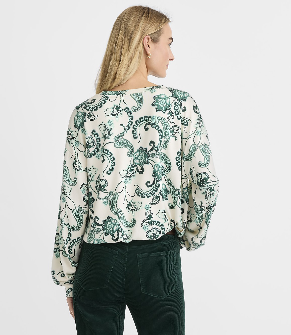 A woman wearing a green and white paisley print blouse and dark green corduroy pants stands against a plain white background.
