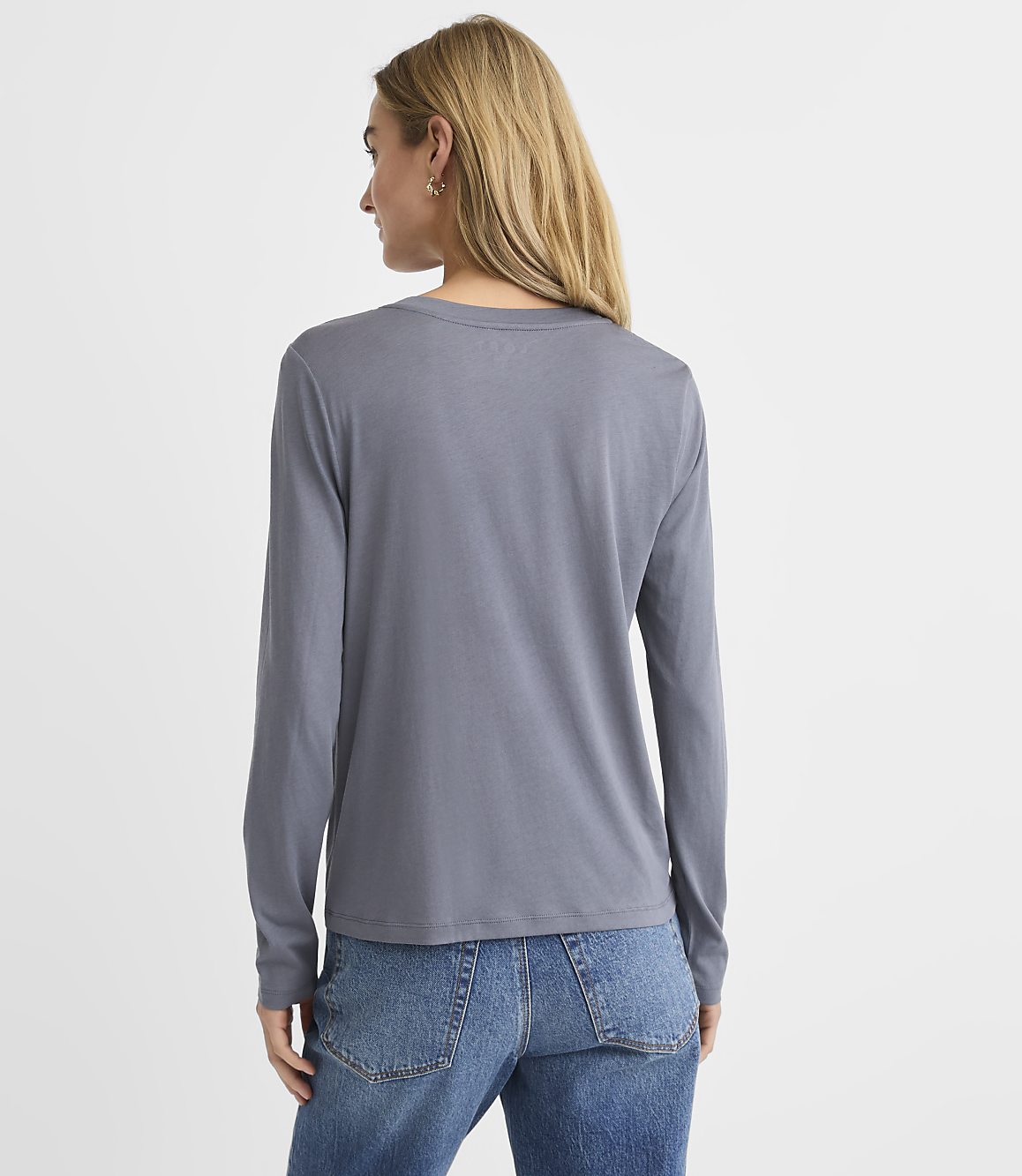 A woman with blonde hair wearing a grey long-sleeved shirt and blue jeans stands against a plain white background.