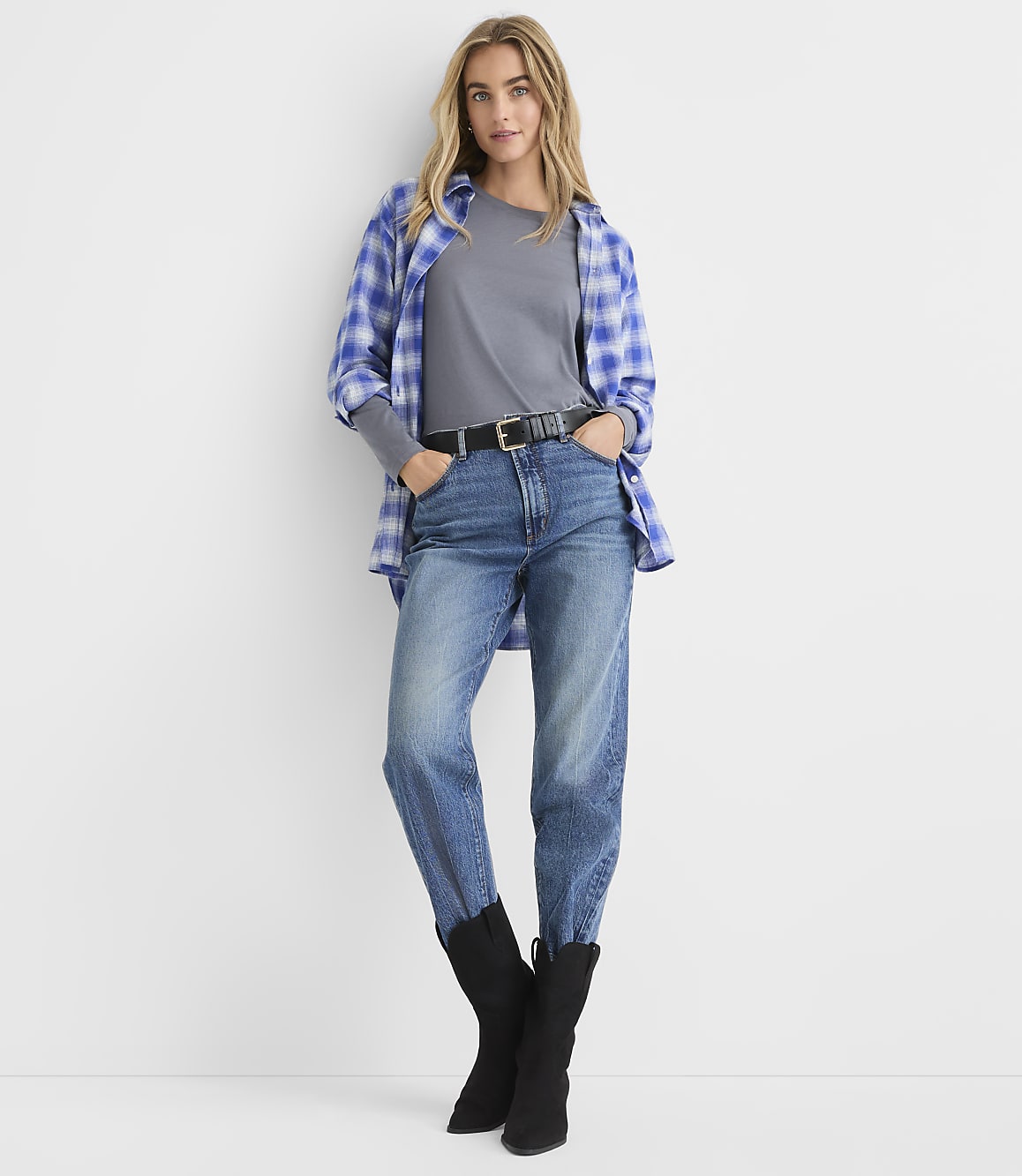 A woman wearing a blue plaid shirt, grey t-shirt, jeans, and black boots stands against a plain white background.