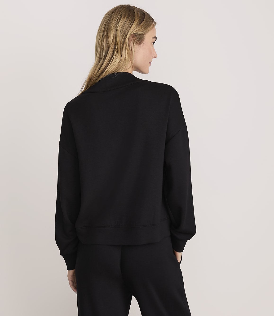 A woman with blonde hair wearing a black sweatshirt and matching pants stands with her back to the camera against a plain white background.