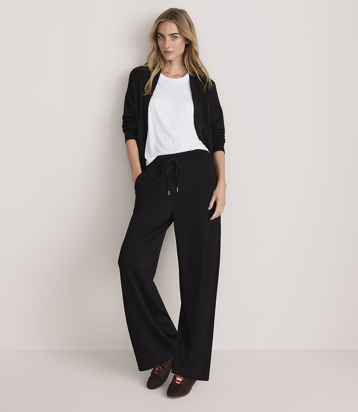 A woman wearing a white t-shirt, black cardigan, and wide-leg black trousers stands against a plain, light-colored background.