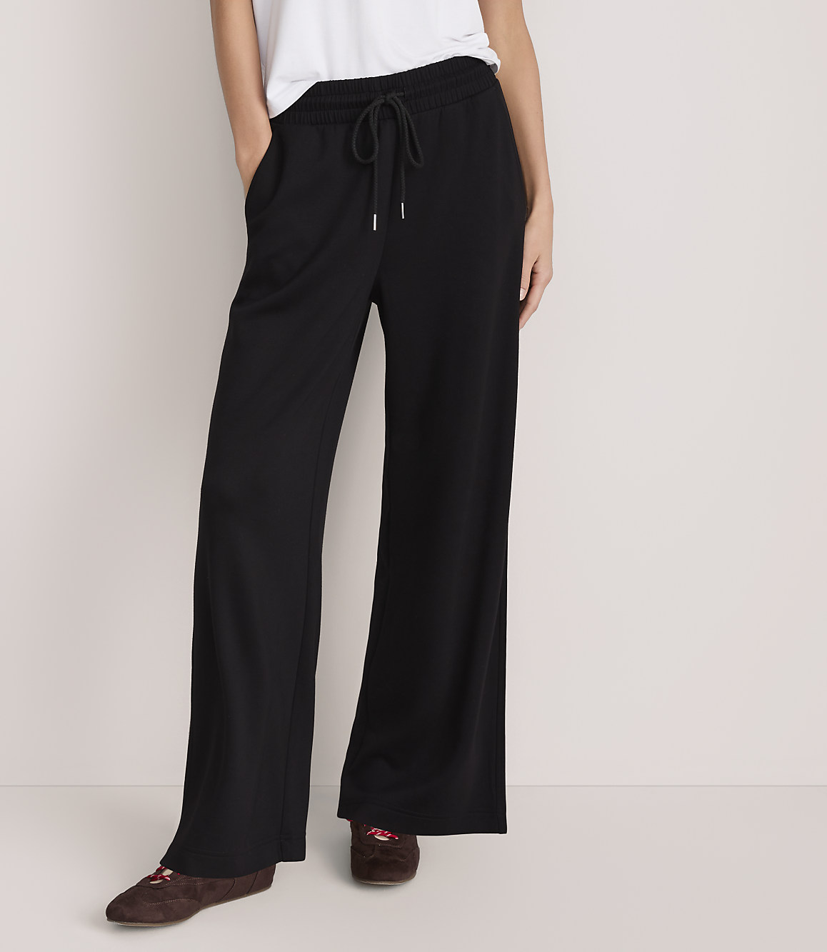 A person wearing a white t-shirt, wide-leg black trousers, and brown shoes stands against a plain, light-colored background.