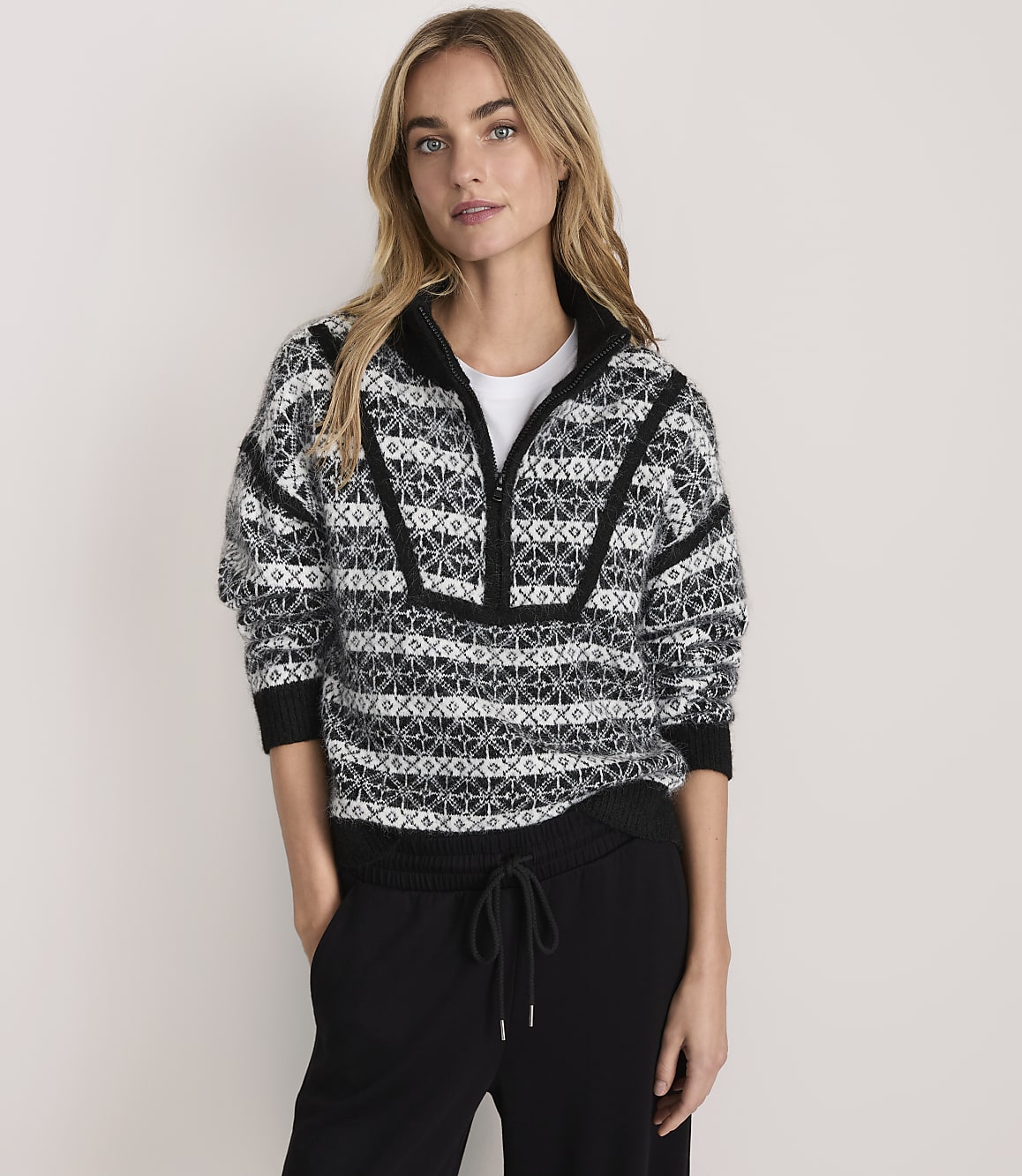A woman wearing a patterned black and white quarter-zip sweater and black pants stands against a plain, light-colored background.