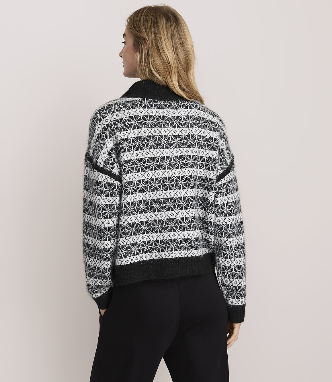 A woman with blonde hair wearing a patterned black and white sweater and black pants stands against a plain light-colored background.
