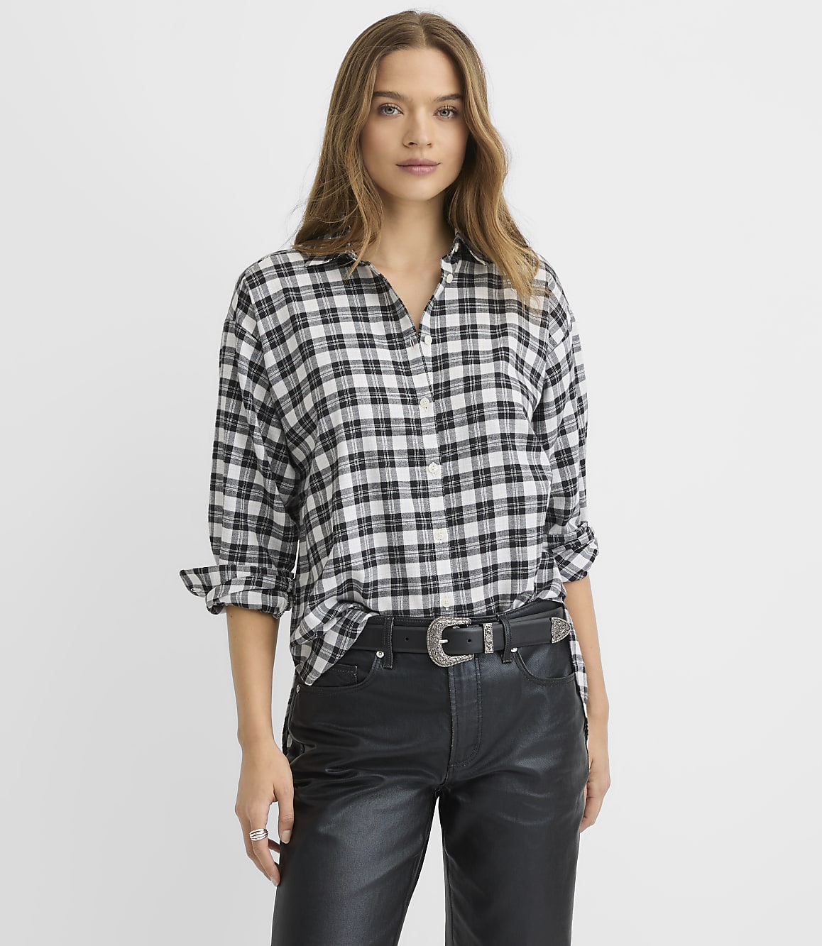 A woman wearing a black and white plaid shirt and black leather pants stands against a plain white background.