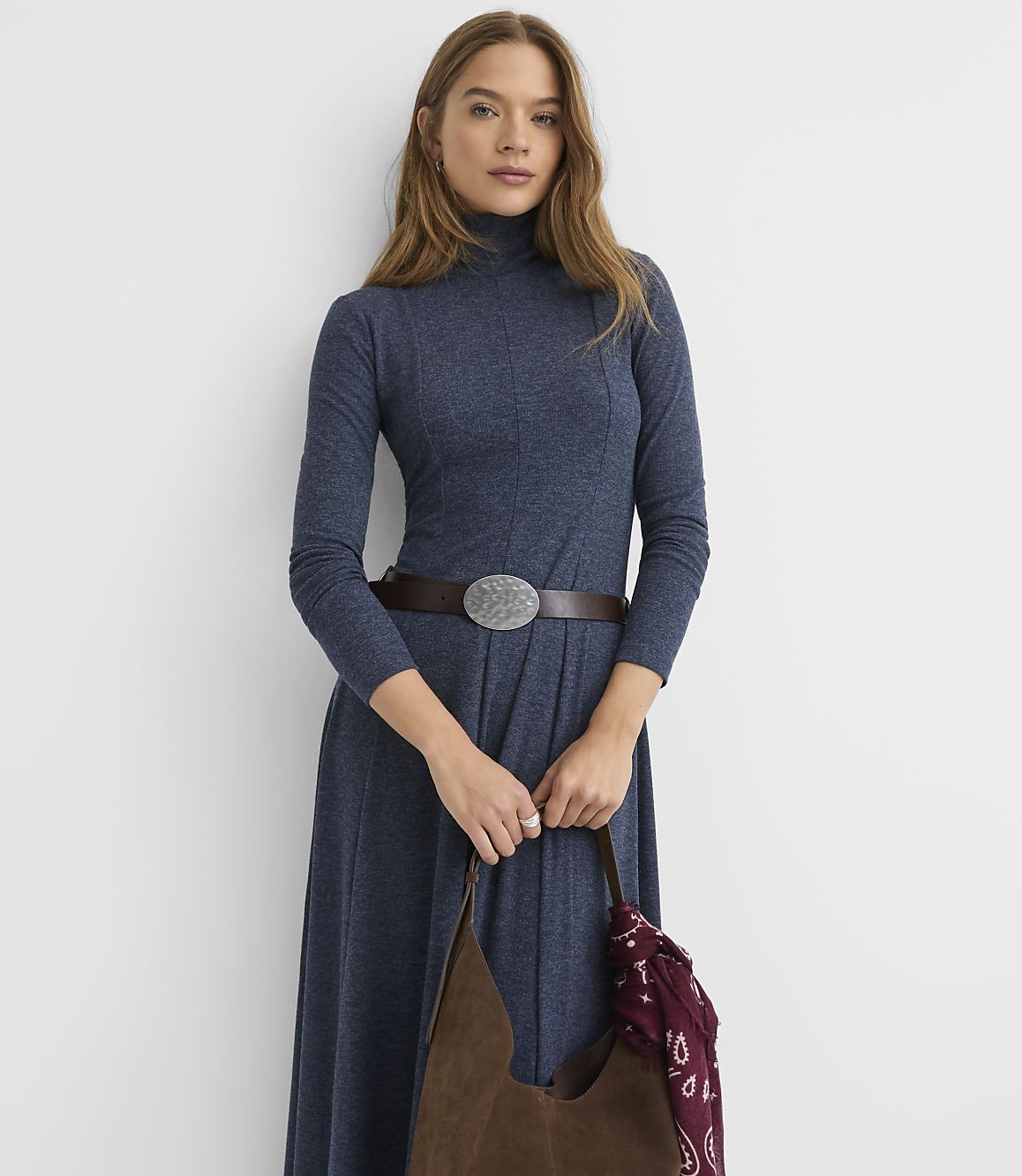 A woman wearing a long-sleeved blue turtleneck dress with a brown belt stands against a plain white background.