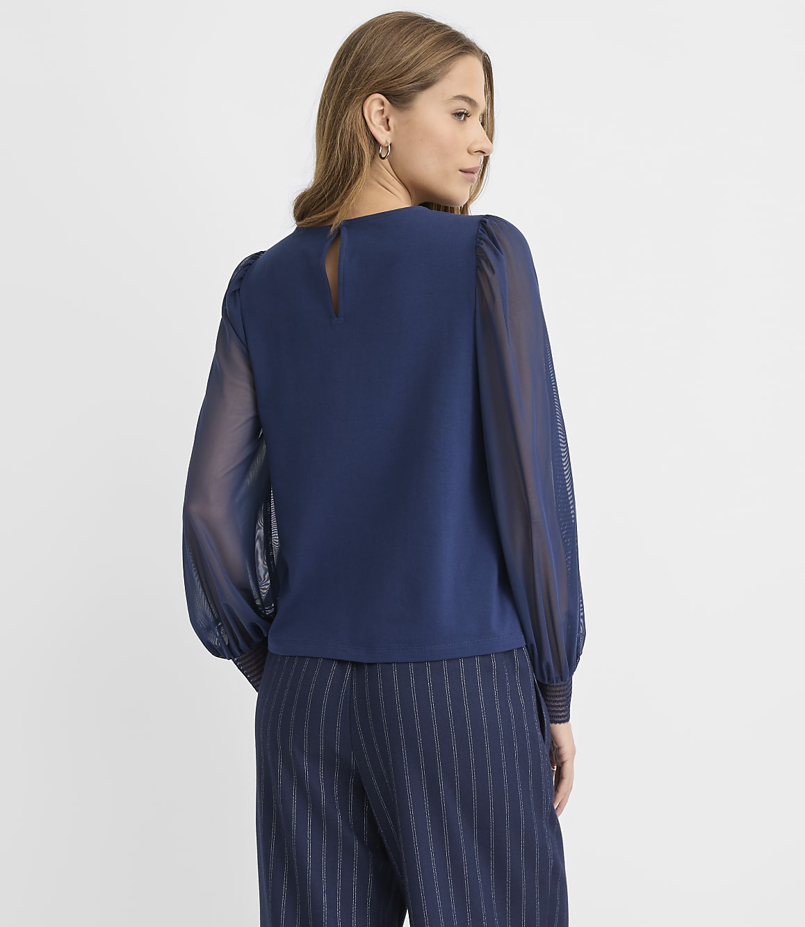 A woman wearing a navy blue blouse with sheer sleeves and pinstriped trousers stands against a plain white background.