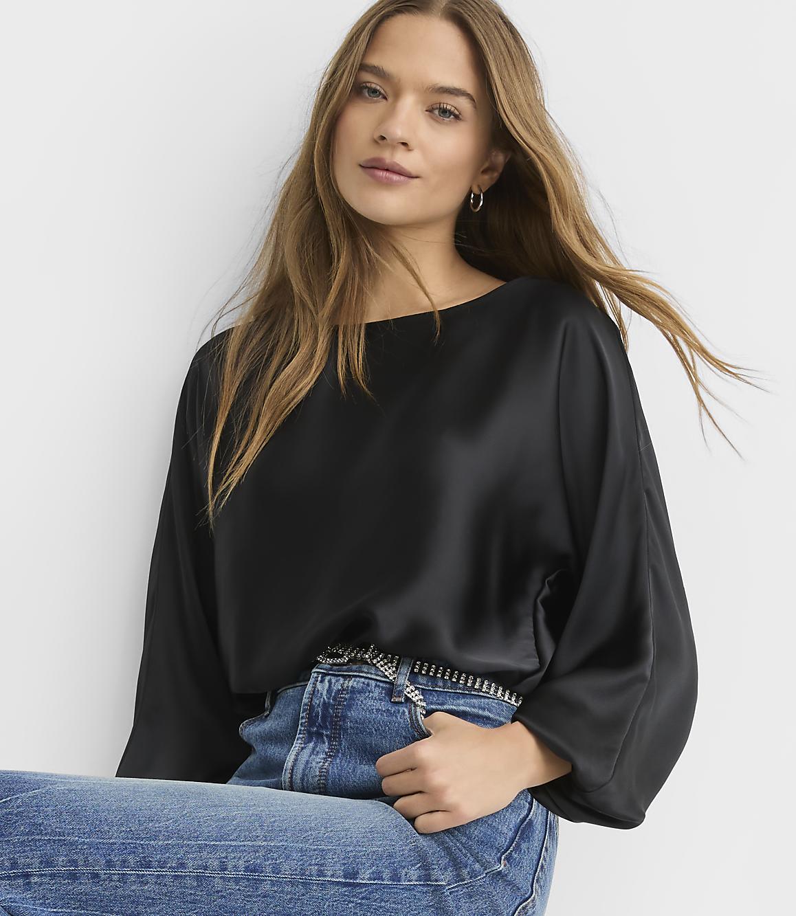 A woman wearing a black satin blouse and blue jeans poses against a plain white background.