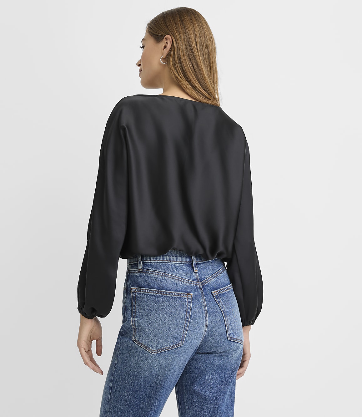 A woman wearing a black satin blouse and blue jeans stands with her back to the camera against a plain white background.