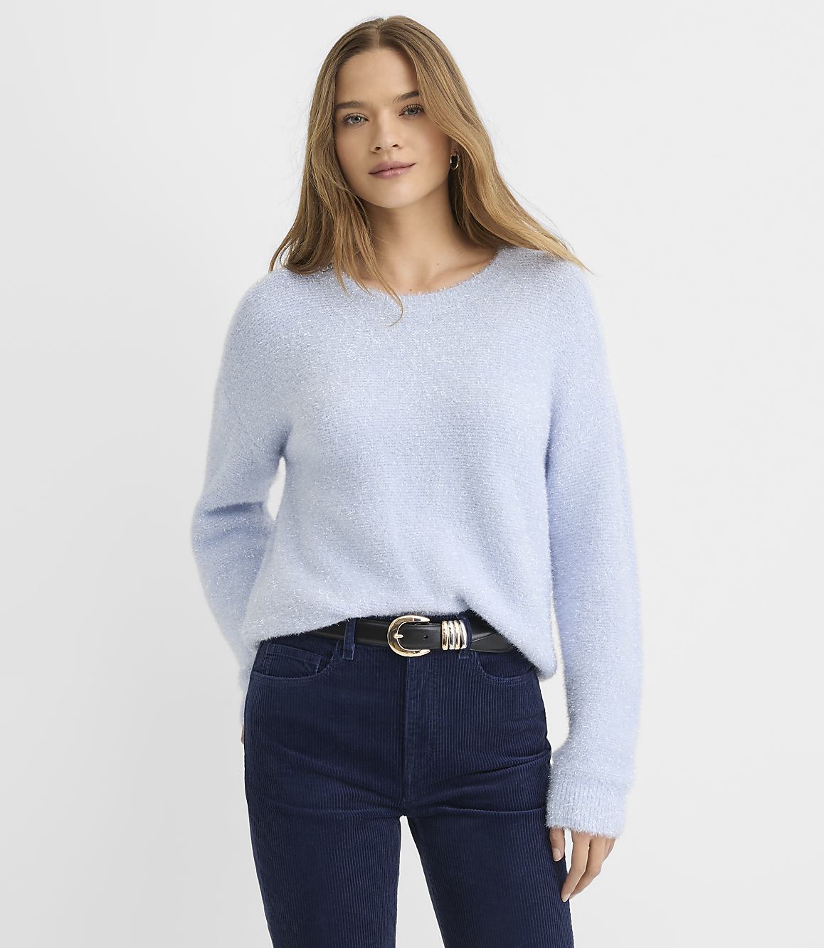 A woman wearing a light blue fuzzy sweater and dark blue corduroy pants stands against a plain white background.