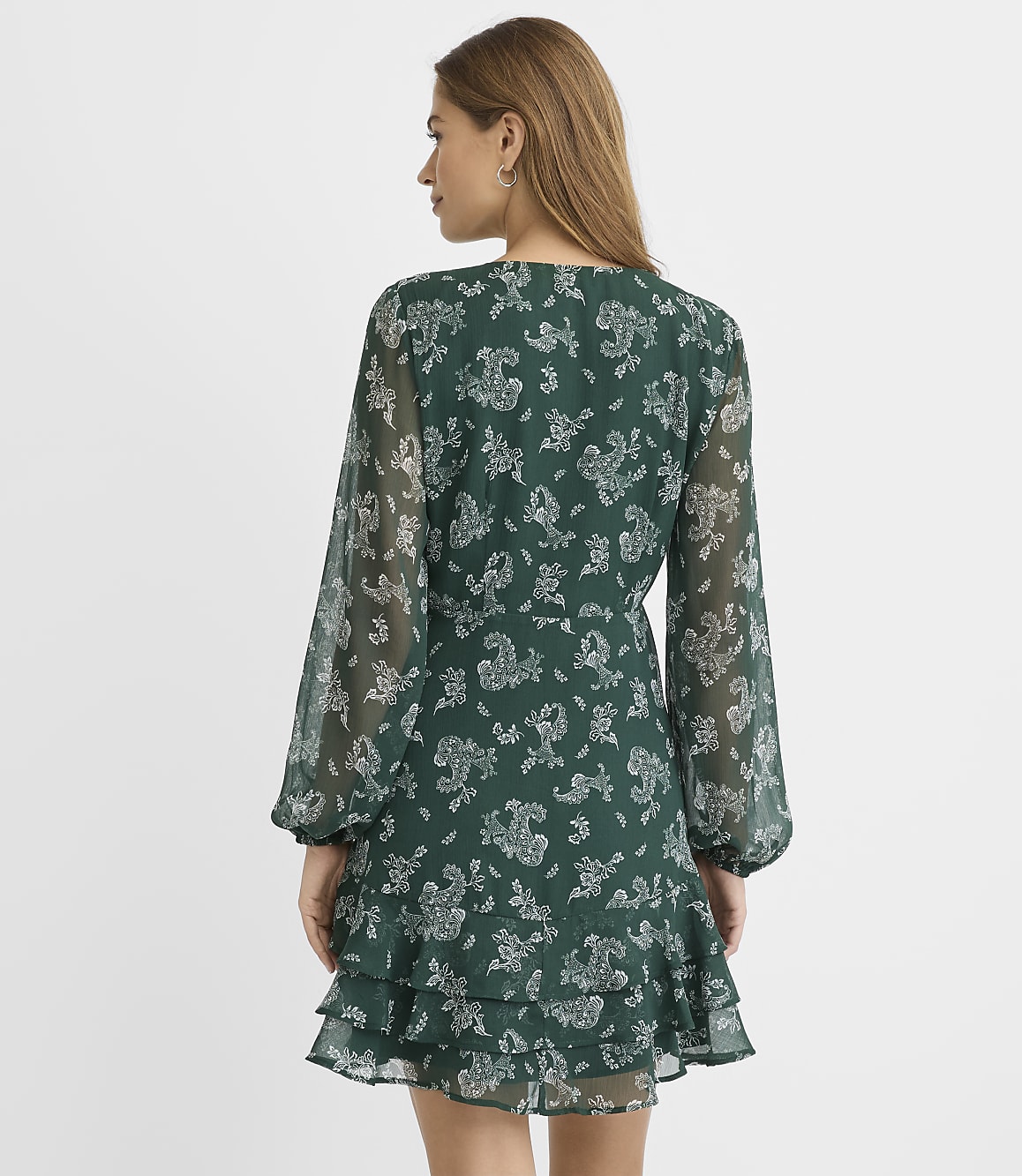 A woman stands with her back to the camera wearing a green floral print dress against a plain white background.