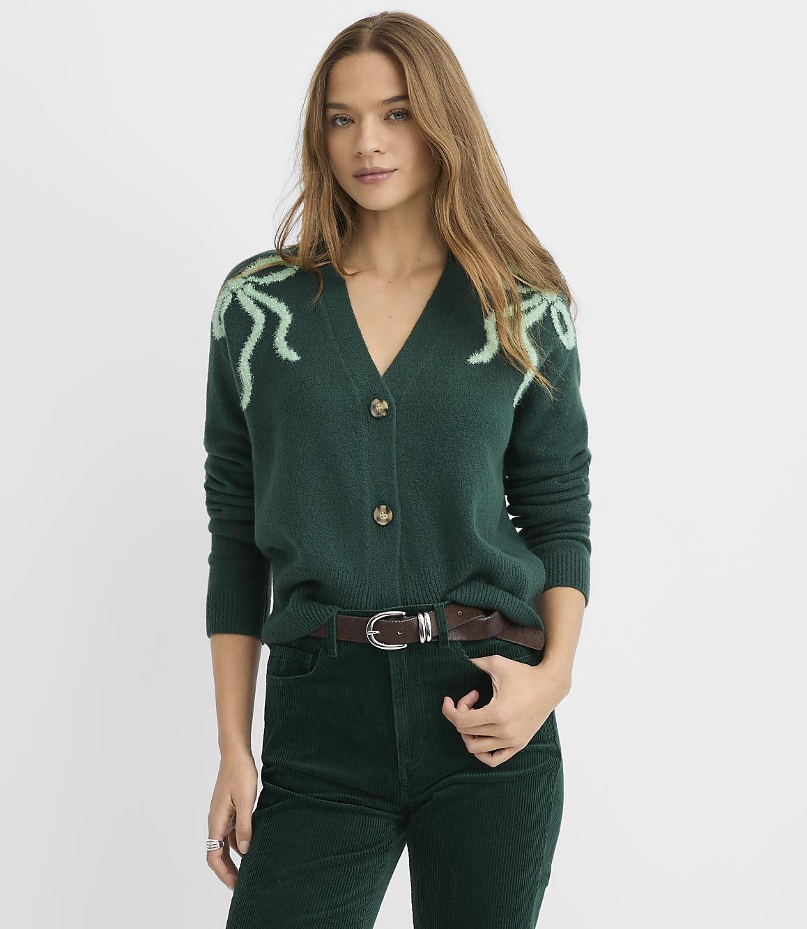 A woman wearing a dark green cardigan with light green accents and matching corduroy pants stands against a plain white background.