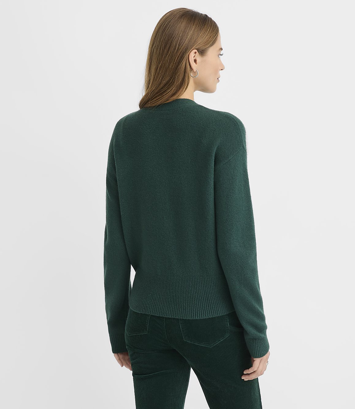 A woman wearing a dark green sweater and matching corduroy pants stands against a plain white background.