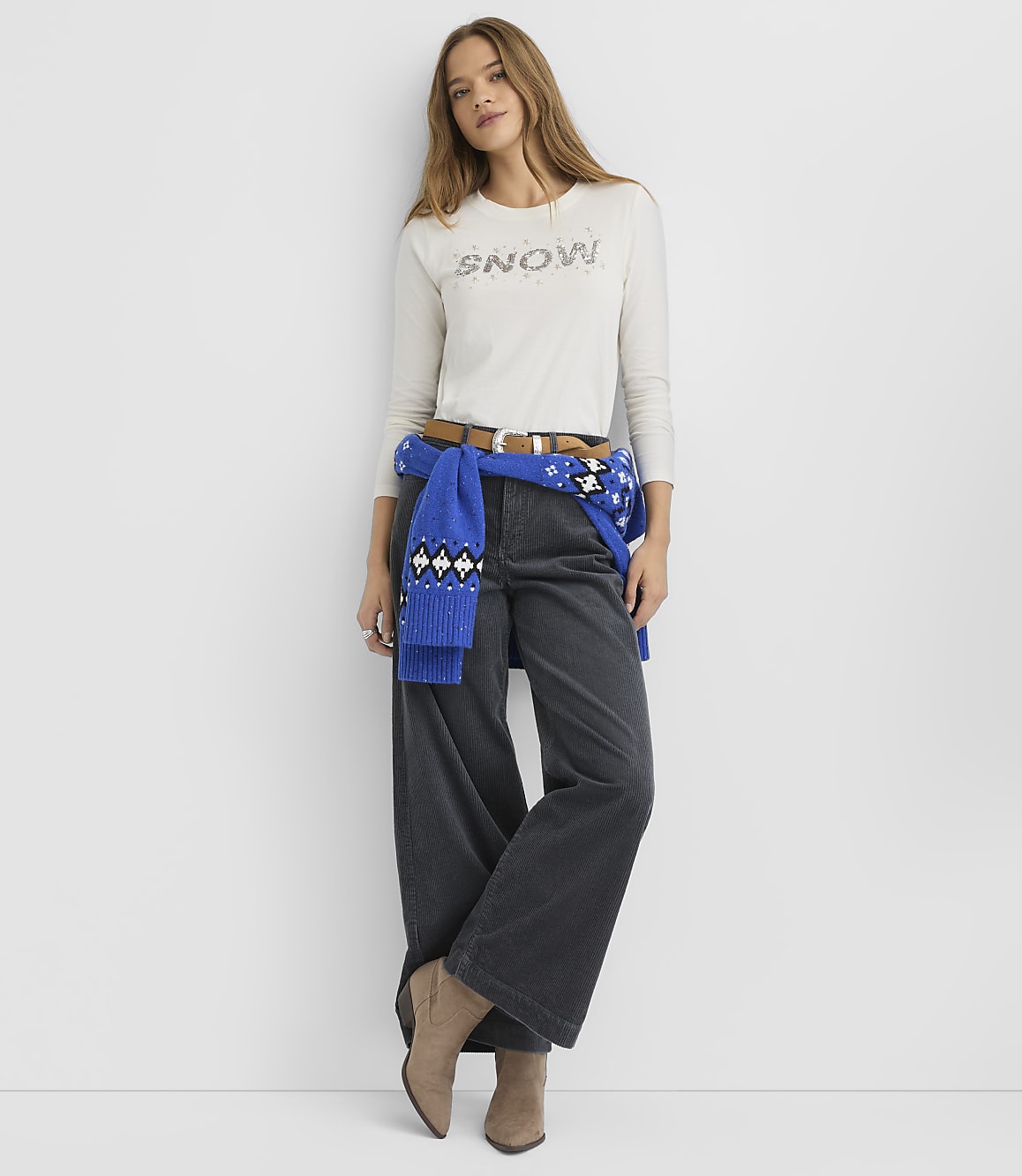 A woman wearing a white "SNOW" shirt, wide-leg dark trousers, and a blue sweater tied around her waist stands against a plain white background.