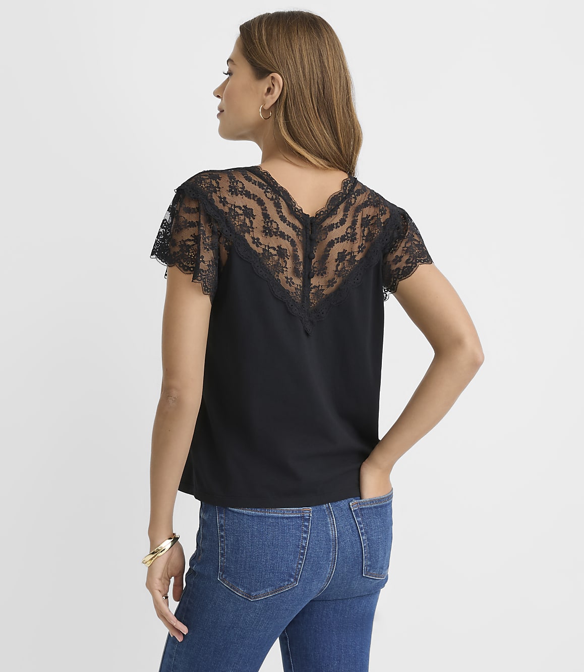 A woman wearing a black lace-detailed top and blue jeans stands against a plain white background.