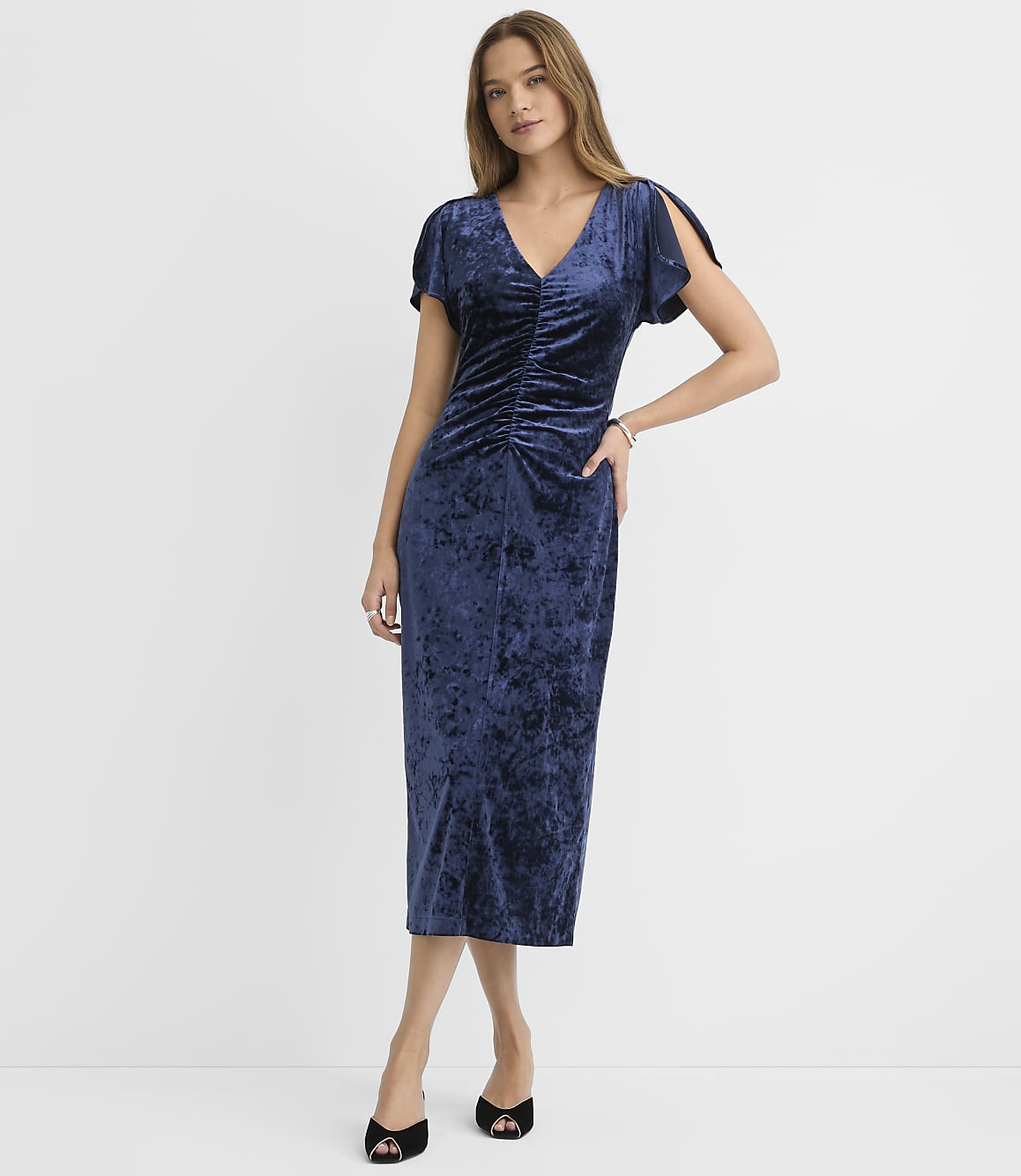 A woman wearing a navy blue velvet midi dress and black heels stands against a plain white background.