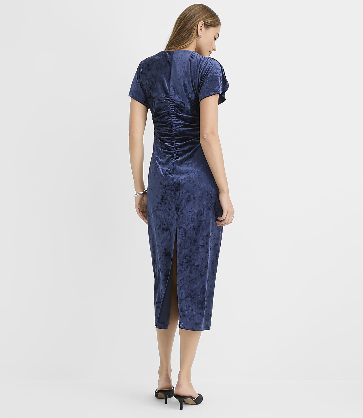 A woman stands with her back to the camera wearing a navy blue velvet midi dress against a plain white background.